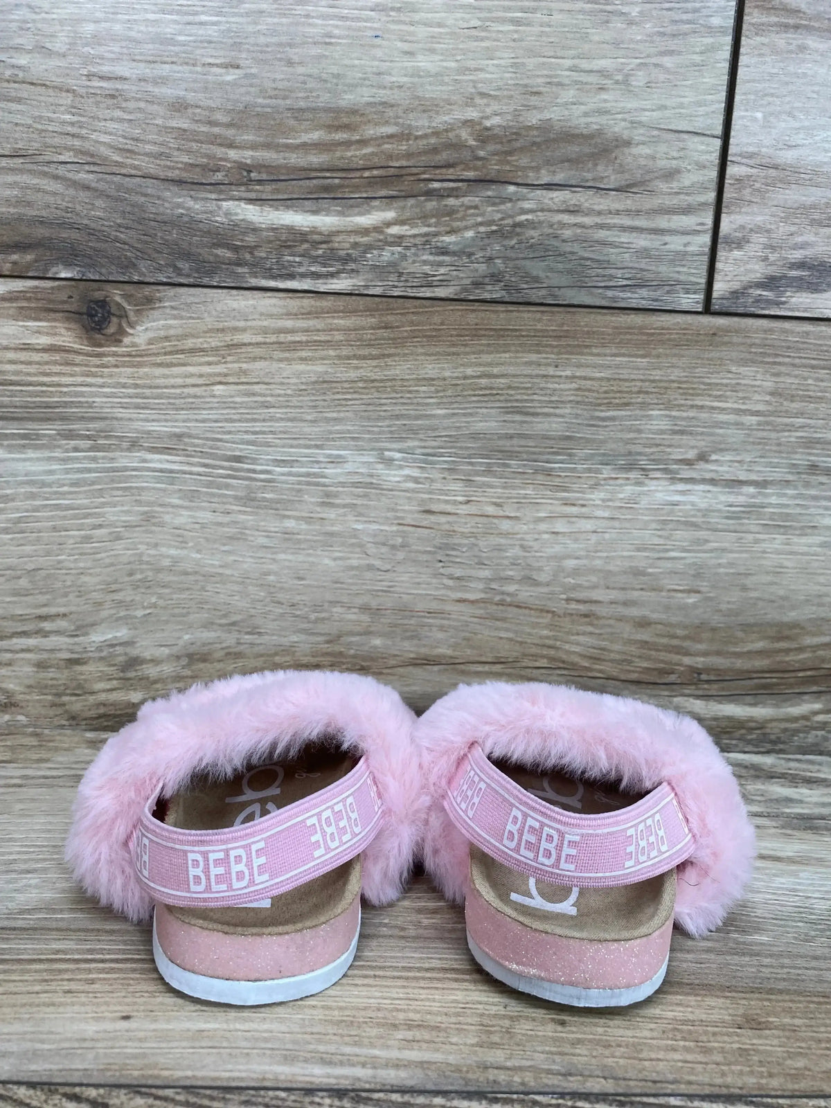 Back view of Bebe pink faux fur sandals showcasing branded logo straps and cork footbeds on a wood plank floor
