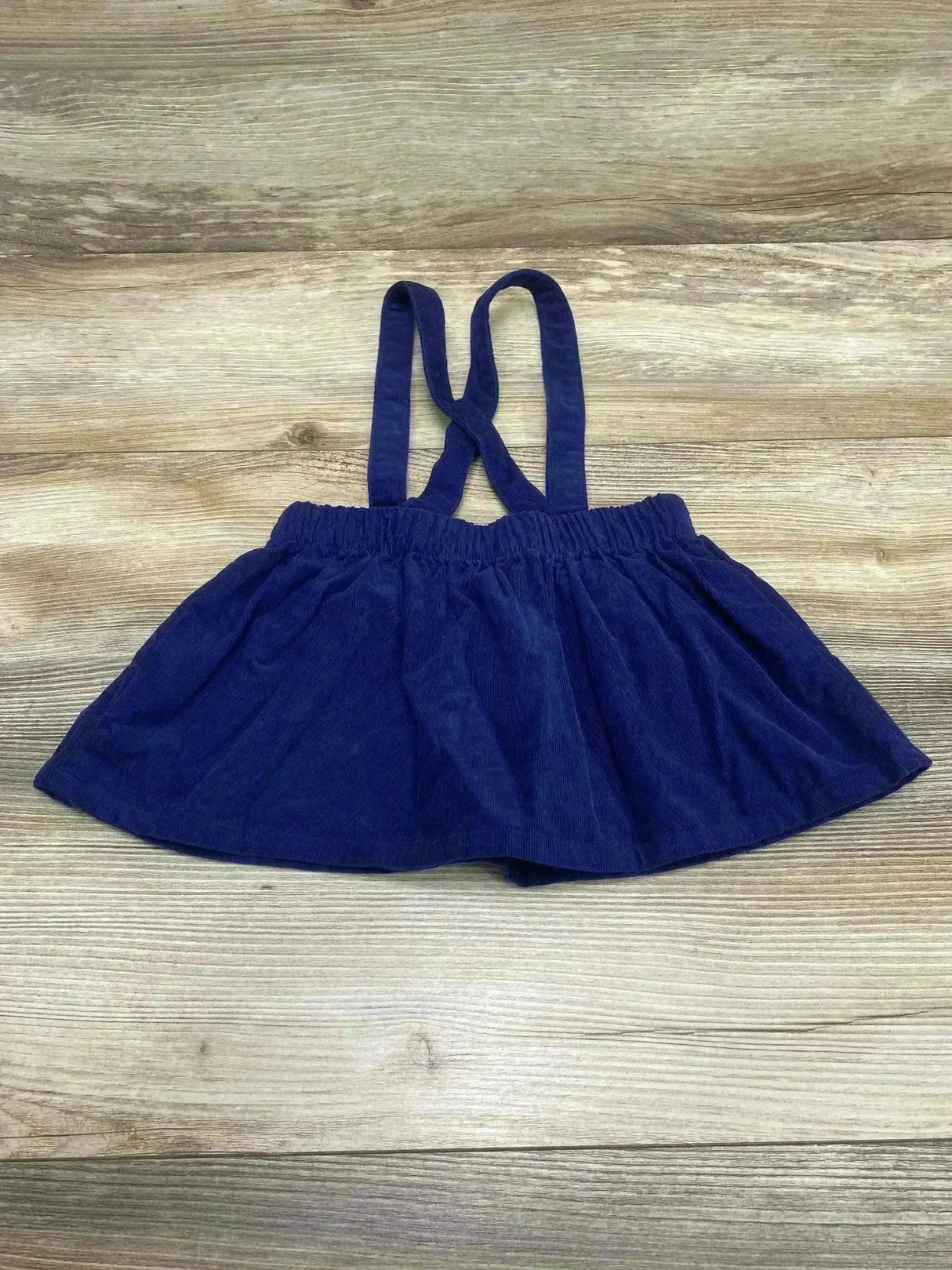 Top down view of navy corduroy suspender skirt with crossed straps, elastic waist and flared hem on wood background
