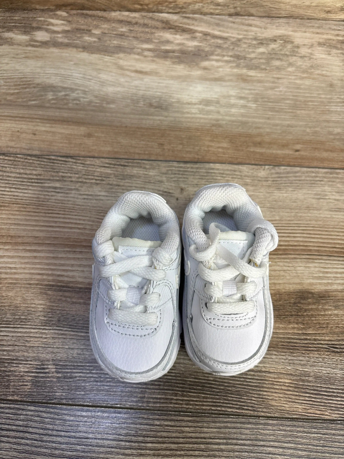 Top view of Nike Air Max 90 TD Triple White sneakers in size 2C showcasing white laces and padded collars on a wood floor