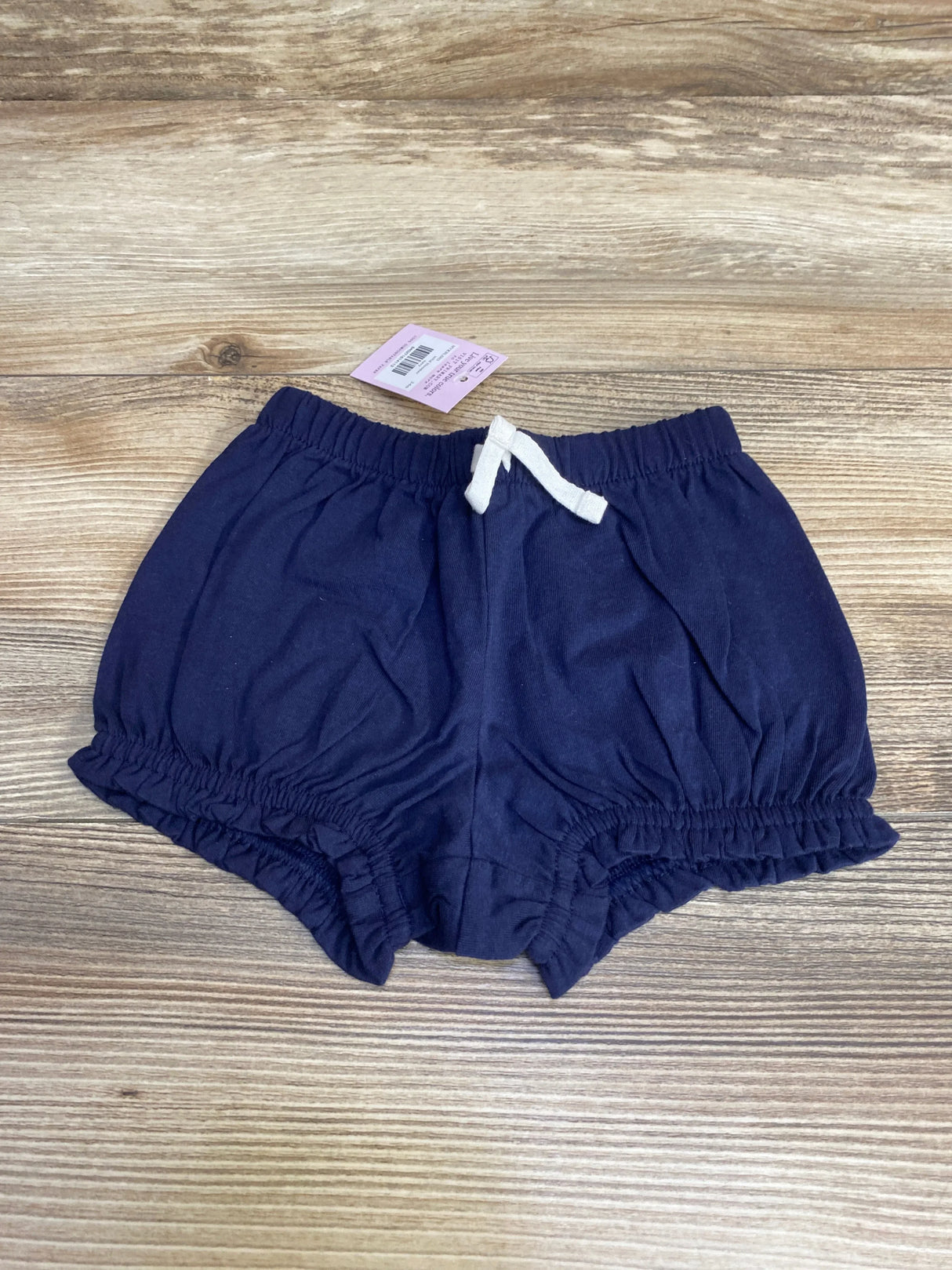 Front view of NWT Primary solid bloomer shorts in navy showcasing white drawstring waist and ruffled leg openings with brand hangtag on wood plank floor
