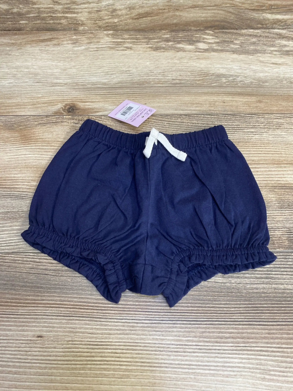 Front view of NWT Primary solid bloomer shorts in navy showcasing white drawstring waist and ruffled leg openings with brand hangtag on wood plank floor
