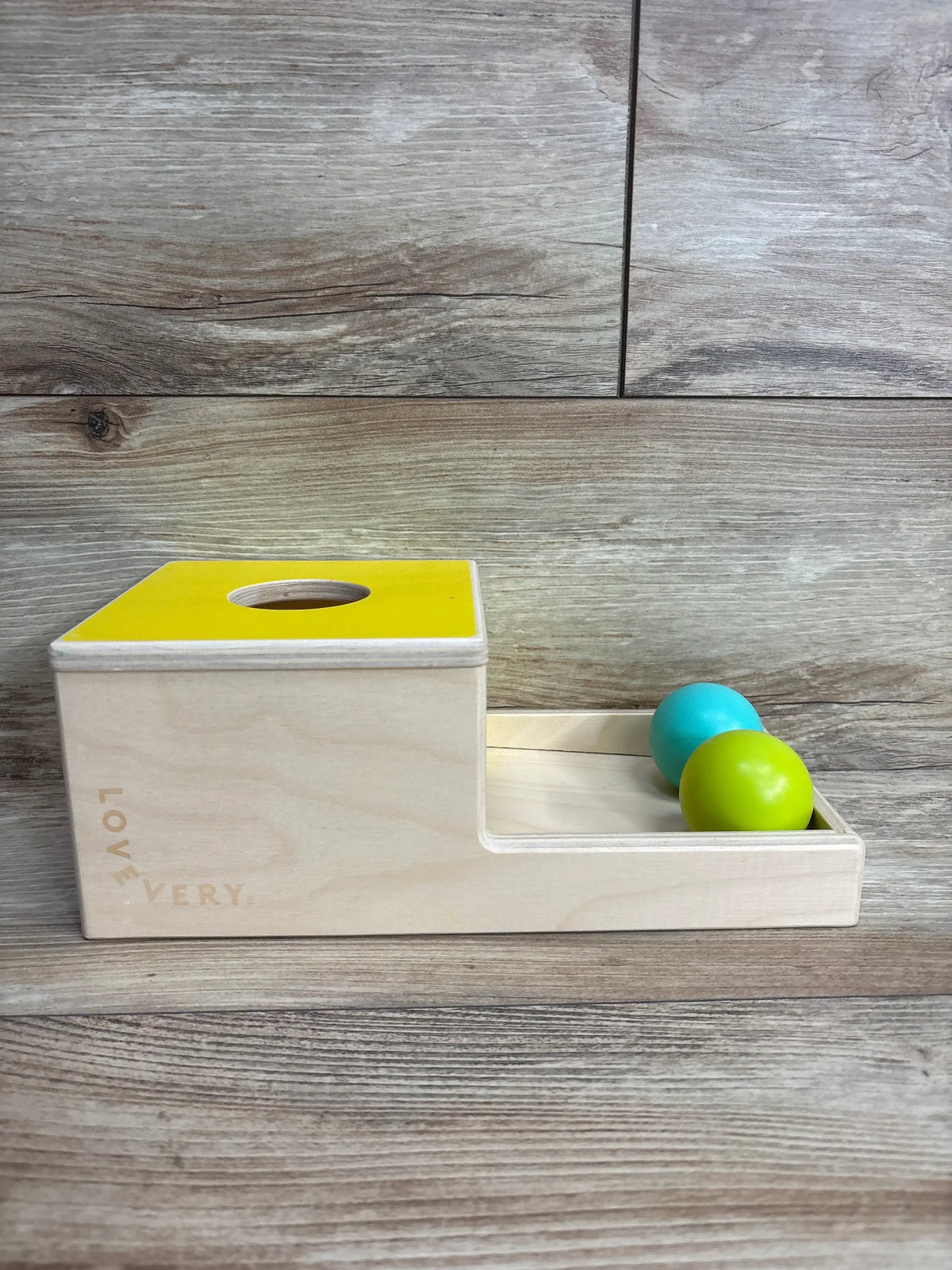 Wooden Montessori ball drop toy with yellow lid, green and blue balls on a wood floor