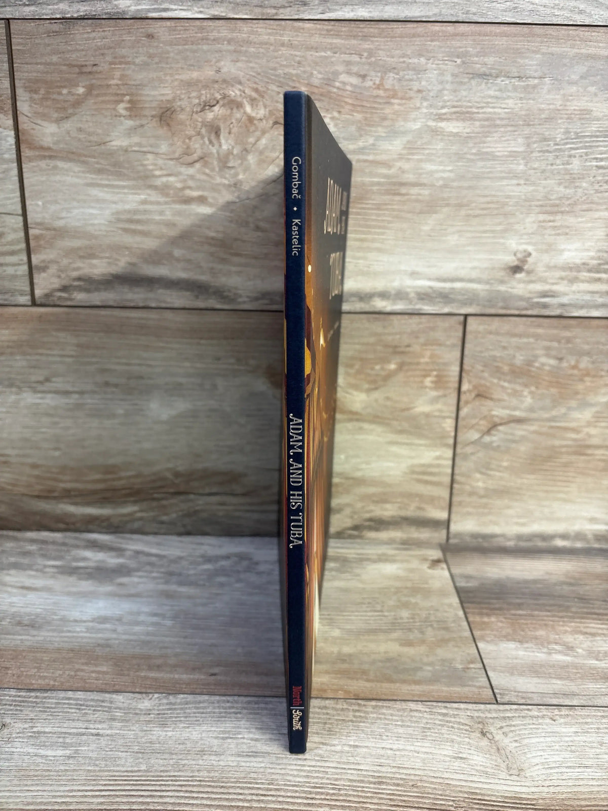 Side view of Adam and His Tuba hardcover book showcasing its title on the spine against a wood-patterned background