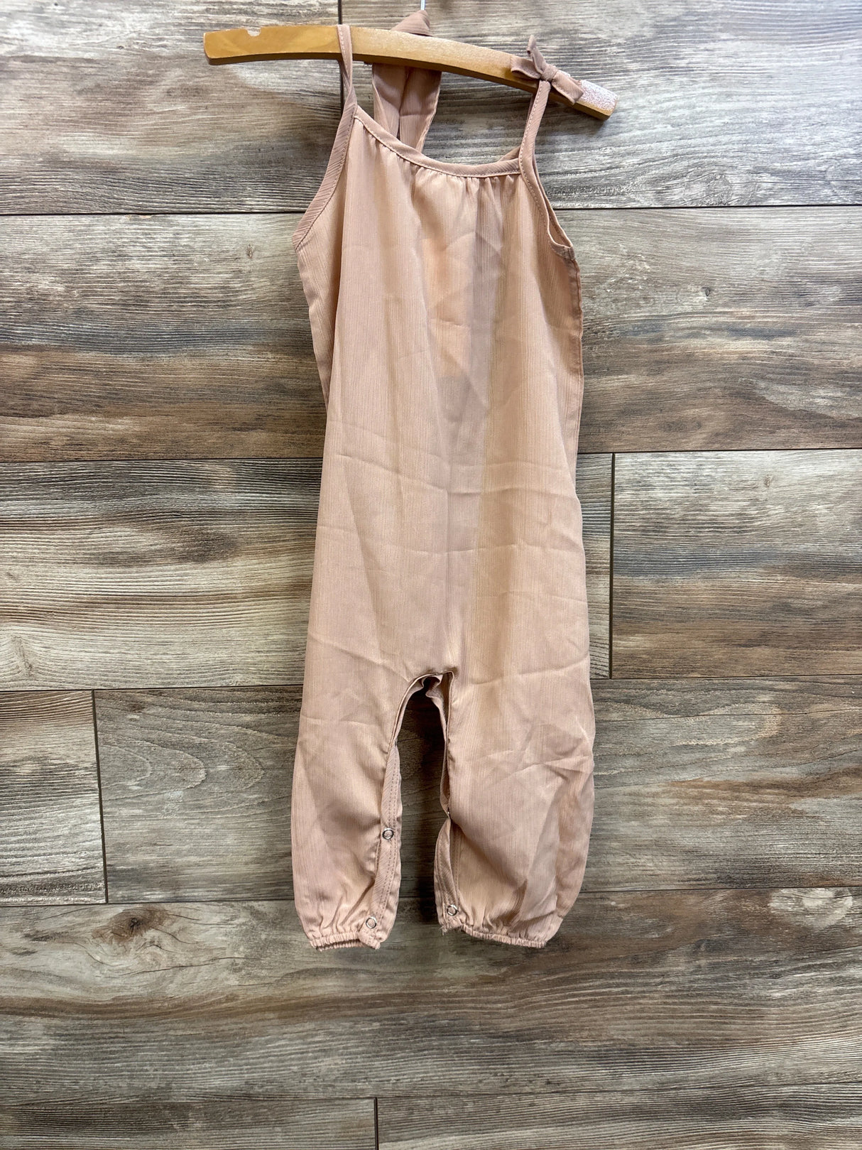 Front view of beige toddler jumpsuit showcasing thin shoulder straps and snap leg closures with wood plank background