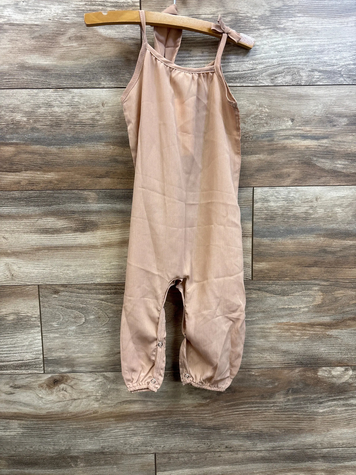 Front view of beige toddler jumpsuit showcasing thin shoulder straps and snap leg closures with wood plank background