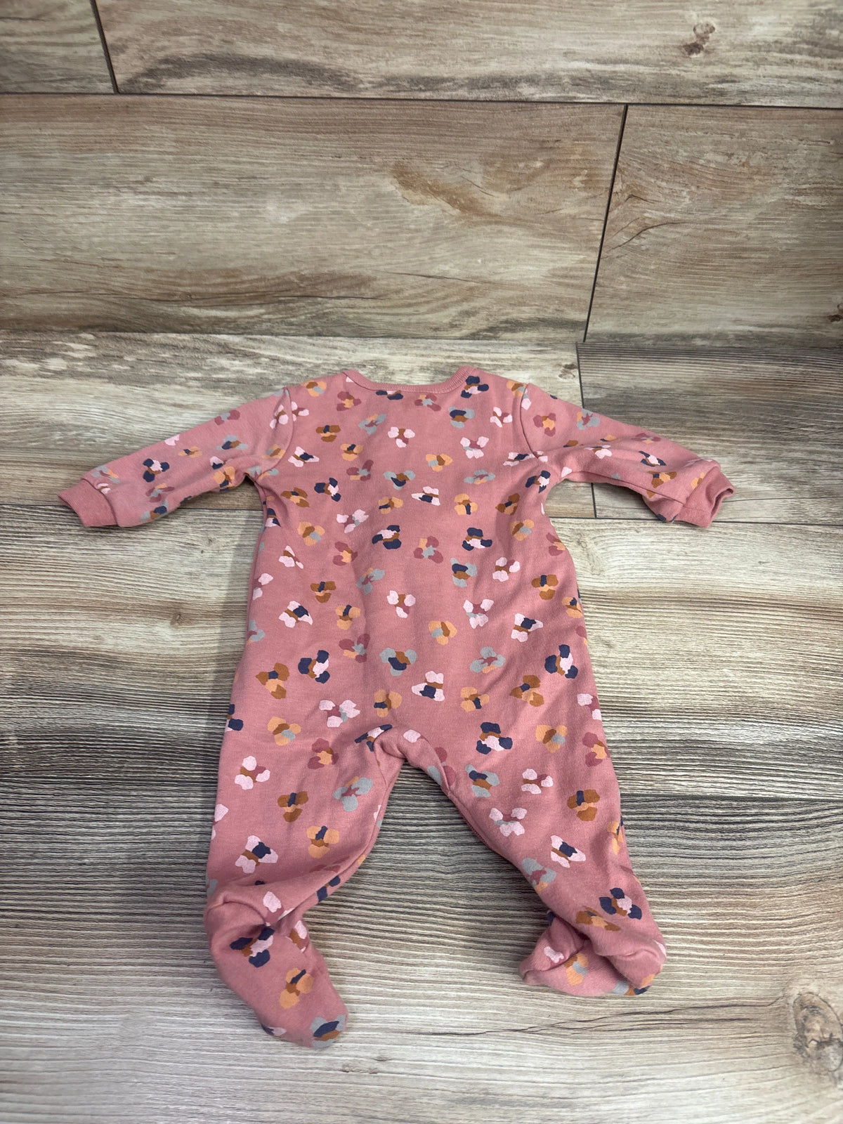Top view of a pink newborn baby sleeper showcasing long sleeves, footed legs, and a multicolored scattered pattern on wood-look flooring