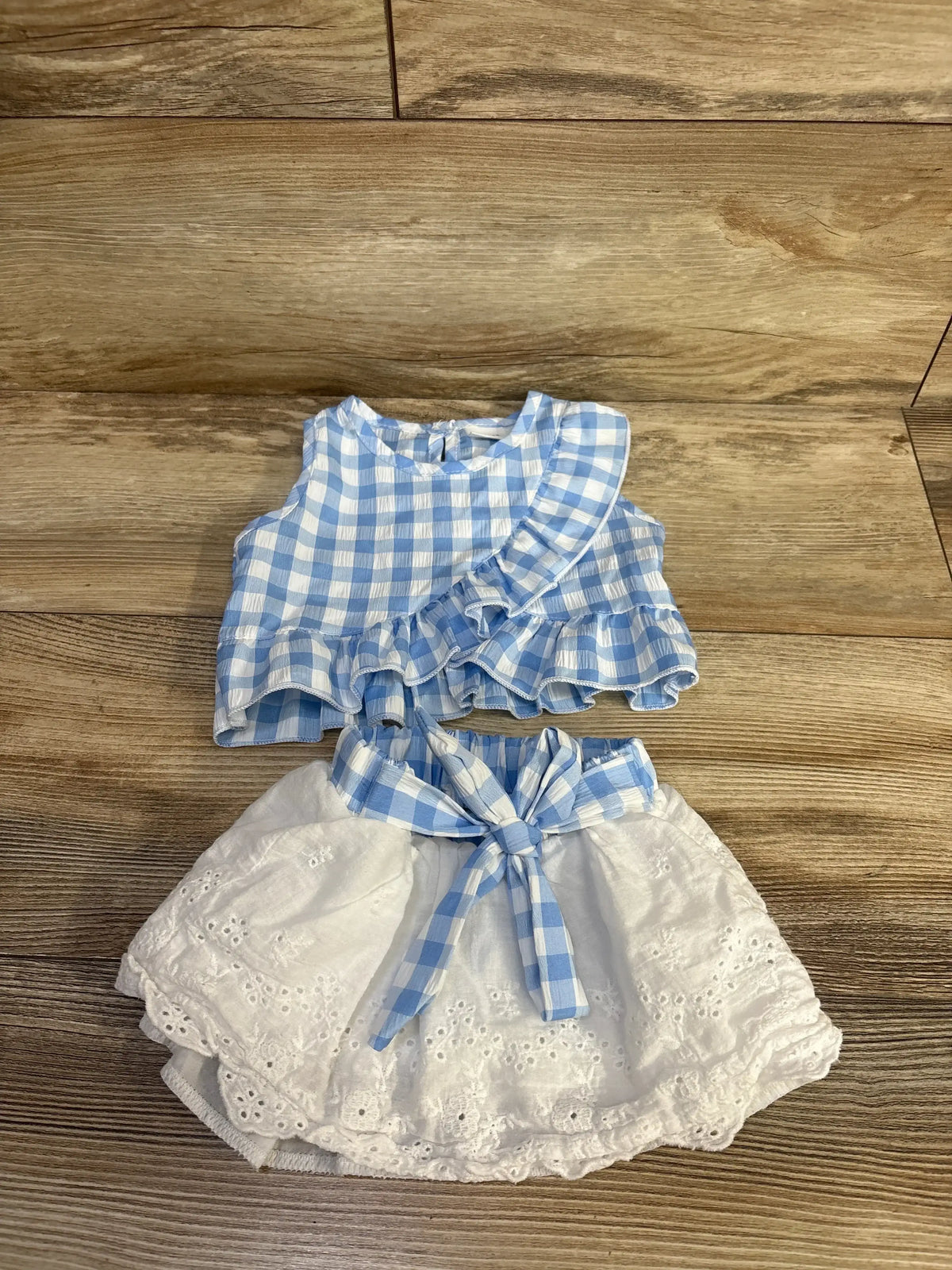 Flat lay of baby outfit: light blue gingham ruffle blouse and white eyelet skirt with gingham tie on wood background