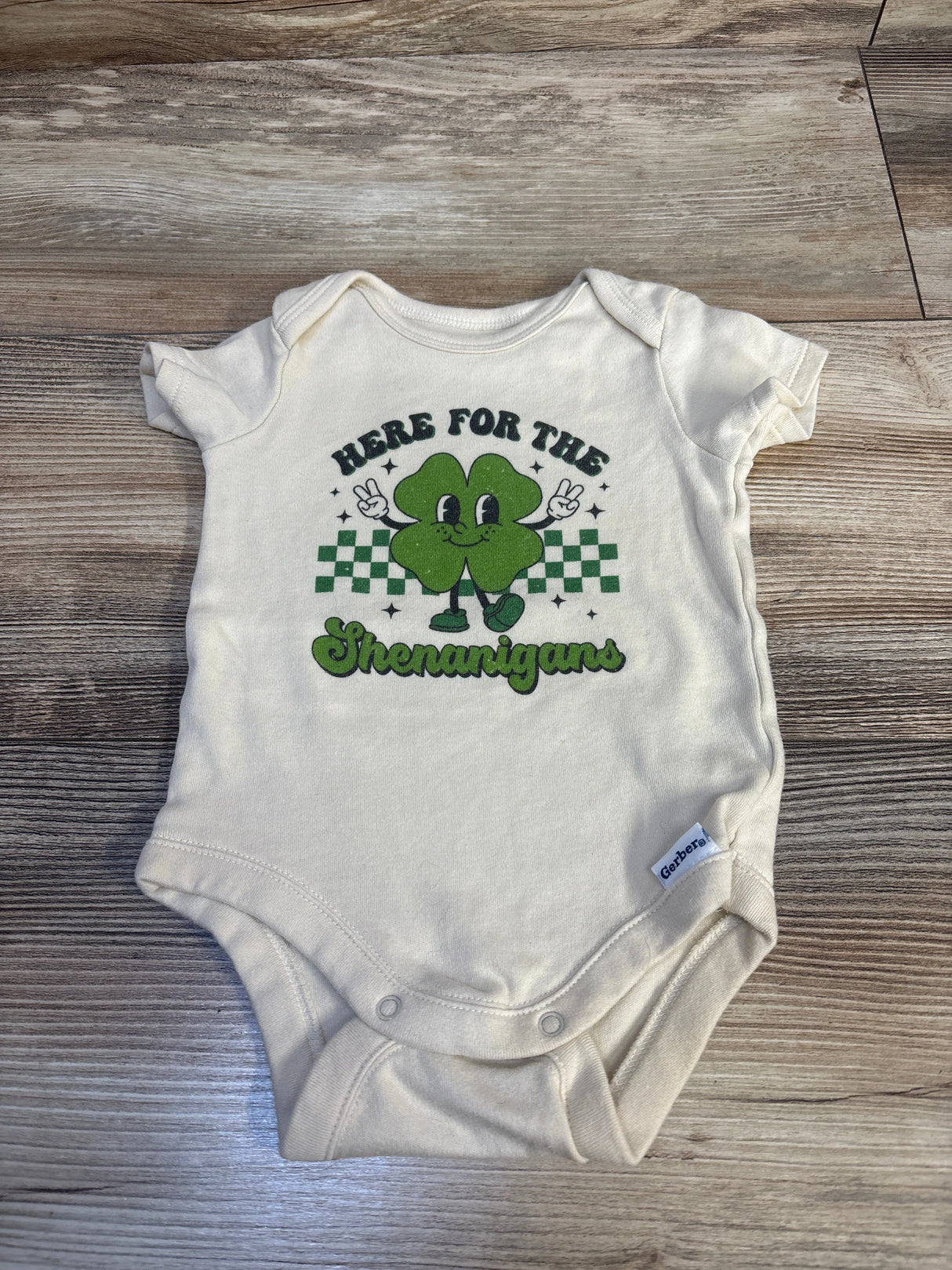 Baby onesie with green shamrock and 'Here for the Shenanigans' text on wood floor