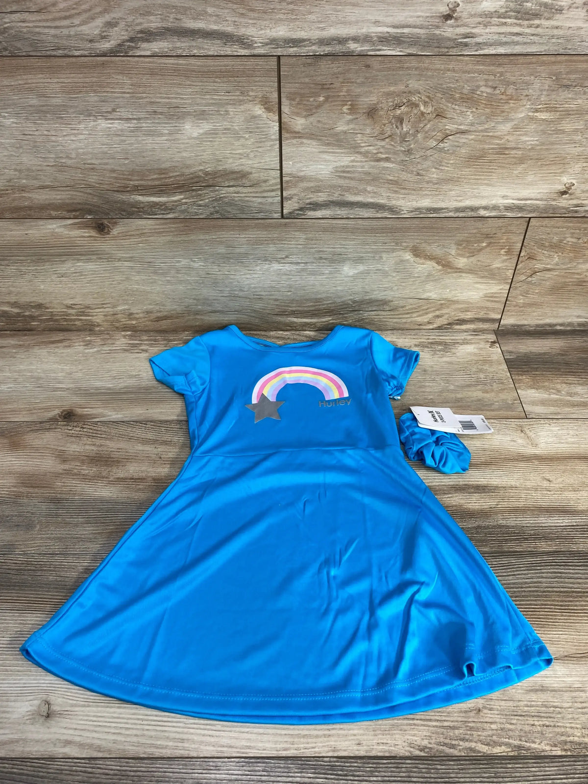 Flat lay of blue Hurley toddler dress and matching scrunchie showcasing front rainbow and star graphic on wood floor