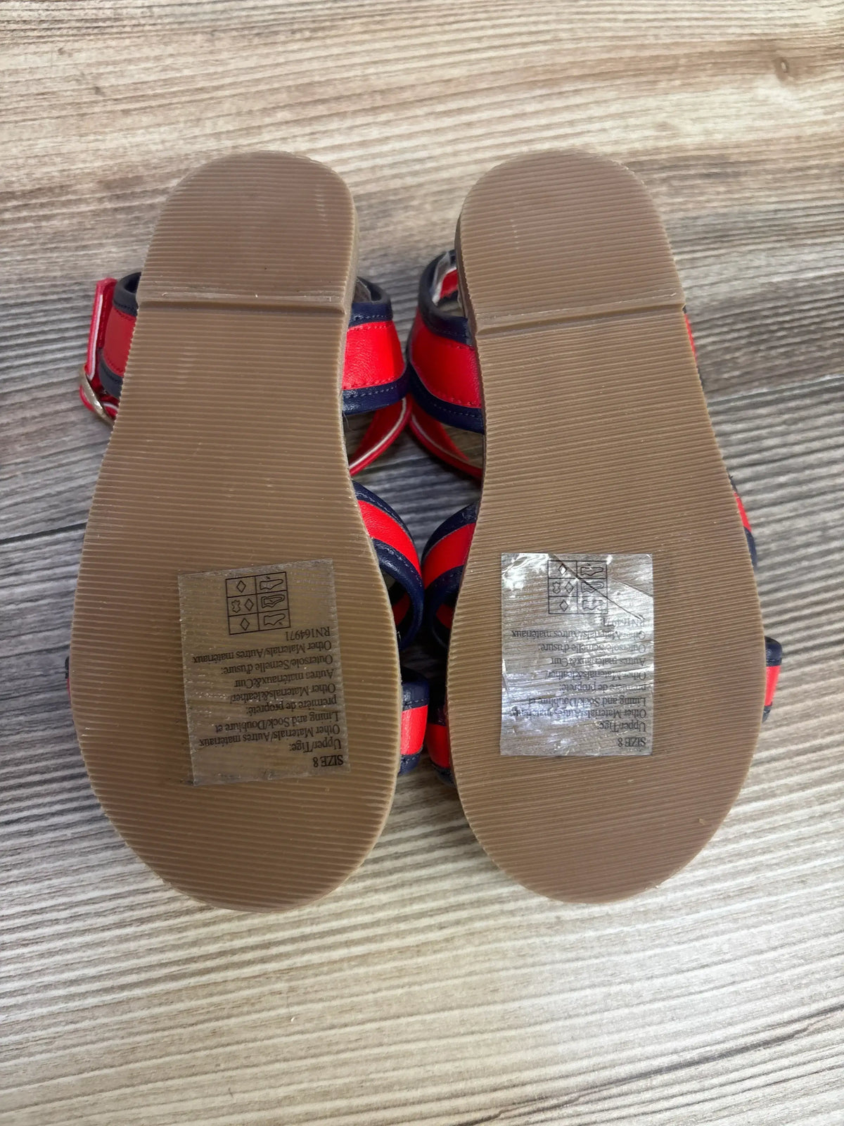 Top-down view of Janie and Jack striped cross-strap toddler sandals showing grooved brown soles and size 8C sticker on wood floor
