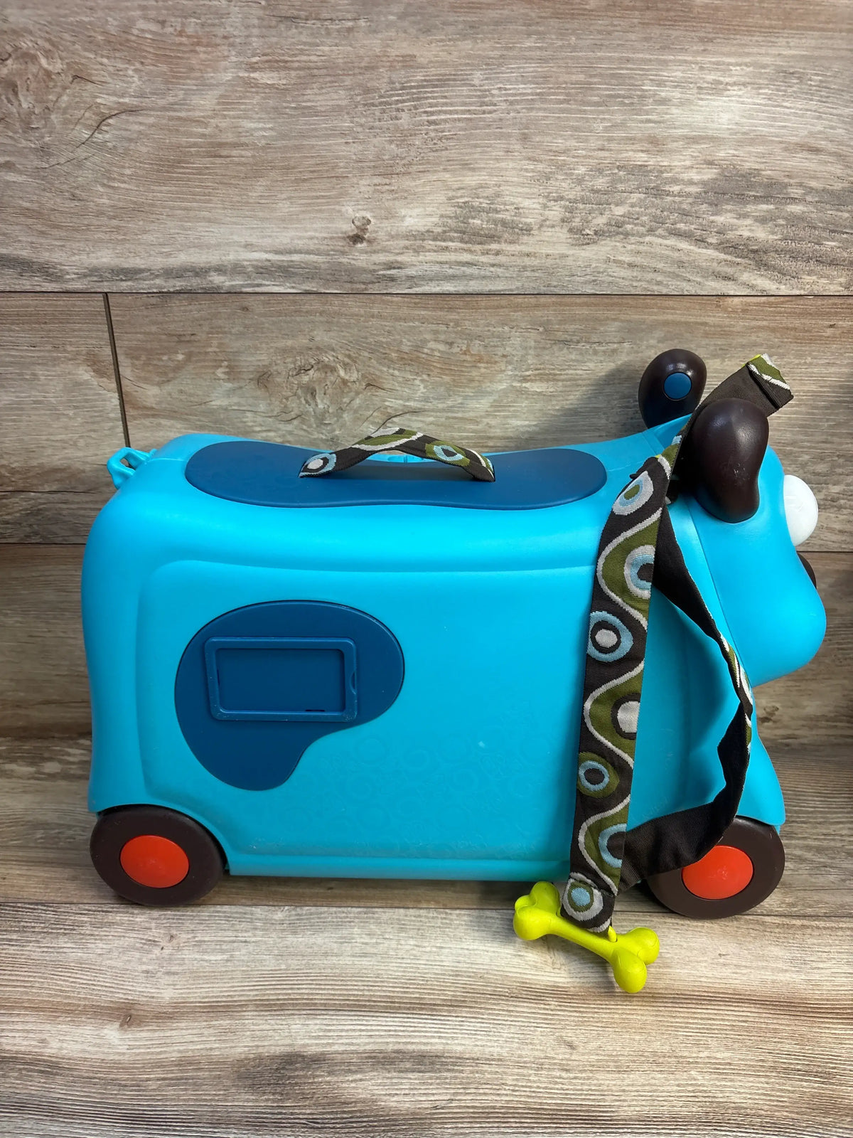 Side view of B. toys On The Go Woofer ride-on suitcase showcasing its turquoise dog design and four wheels on a wood-look floor