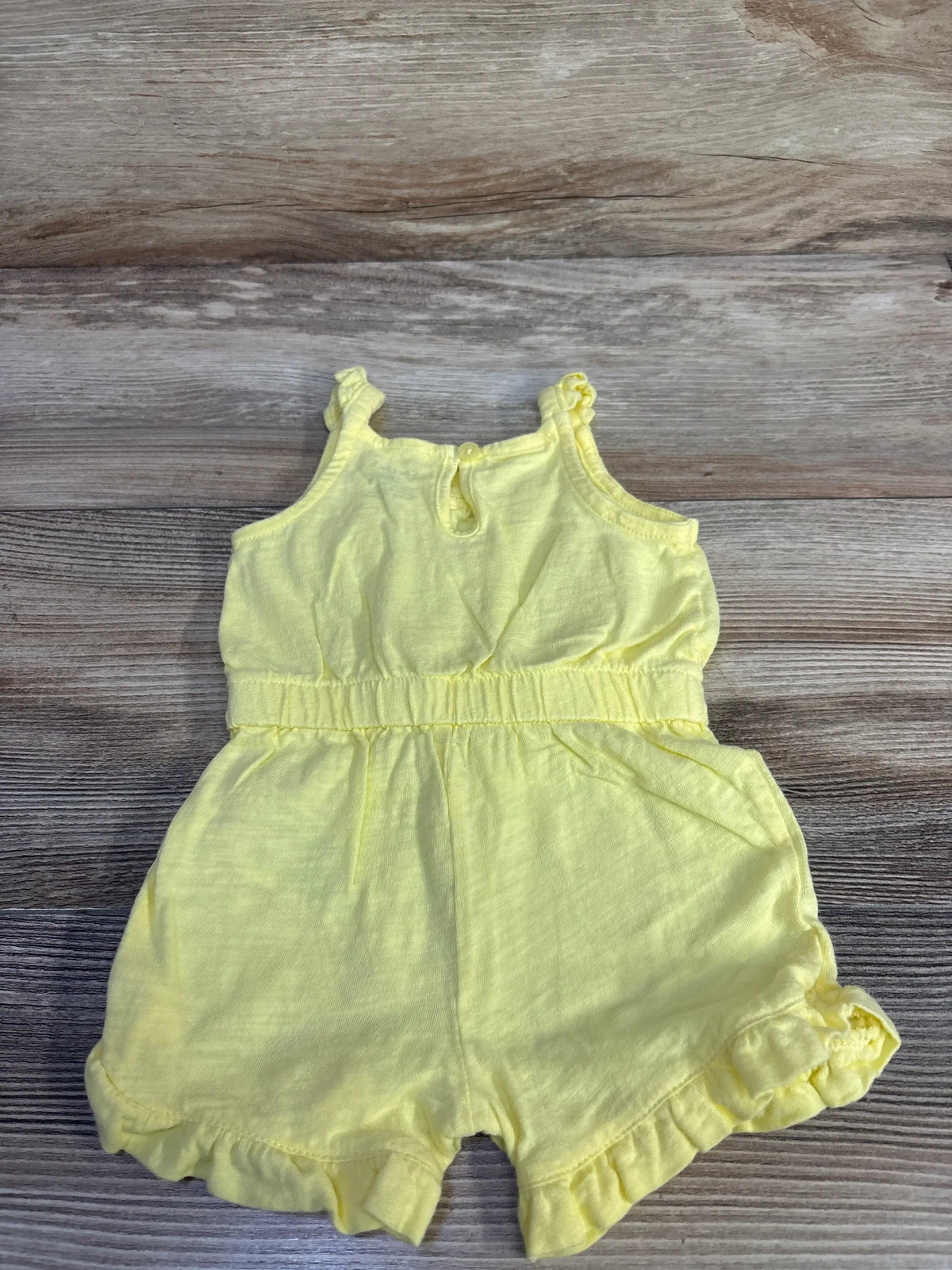 Flat-lay view of yellow sleeveless shortie romper showing elastic waist and ruffle leg trim on wood background