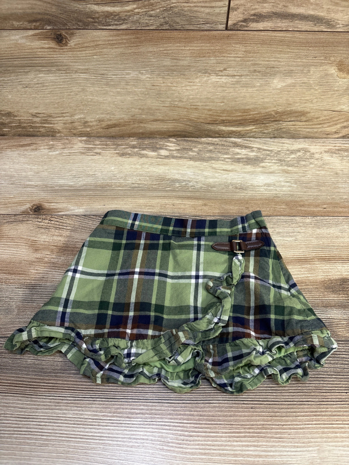 Front view of Janie & Jack The Equestrian plaid skirt in green with a ruffled hem and belt detail on wood