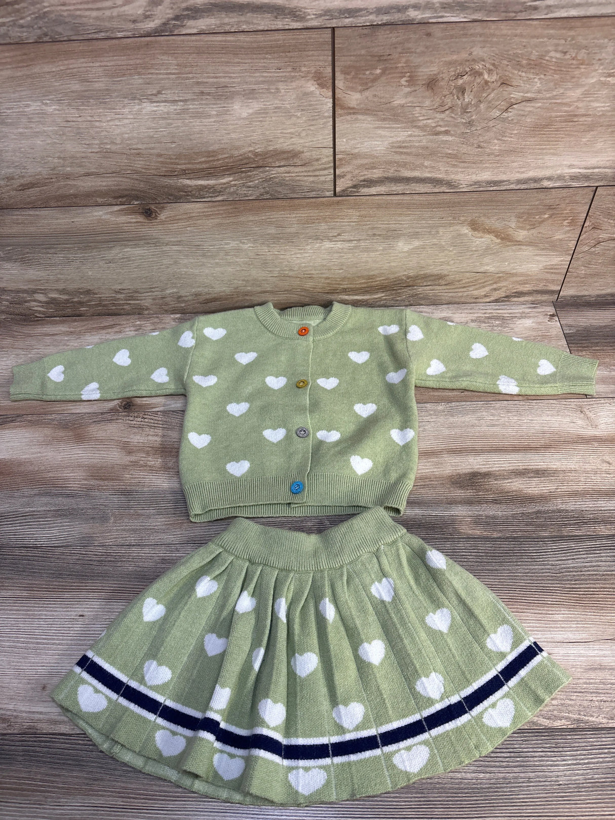 Green knit sweater and pleated skirt set with white heart patterns and colorful buttons on wooden floor
