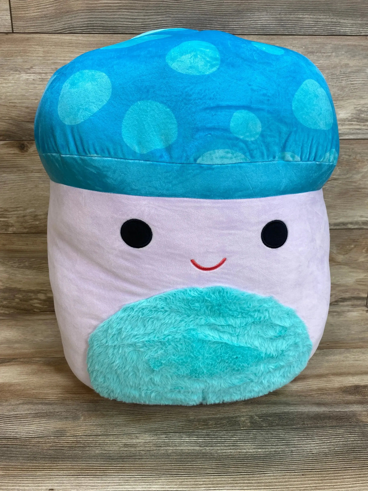Front view of 20in Squishmallows Pyle mushroom plush with blue polka-dot cap, pink body and teal fuzzy belly on wood