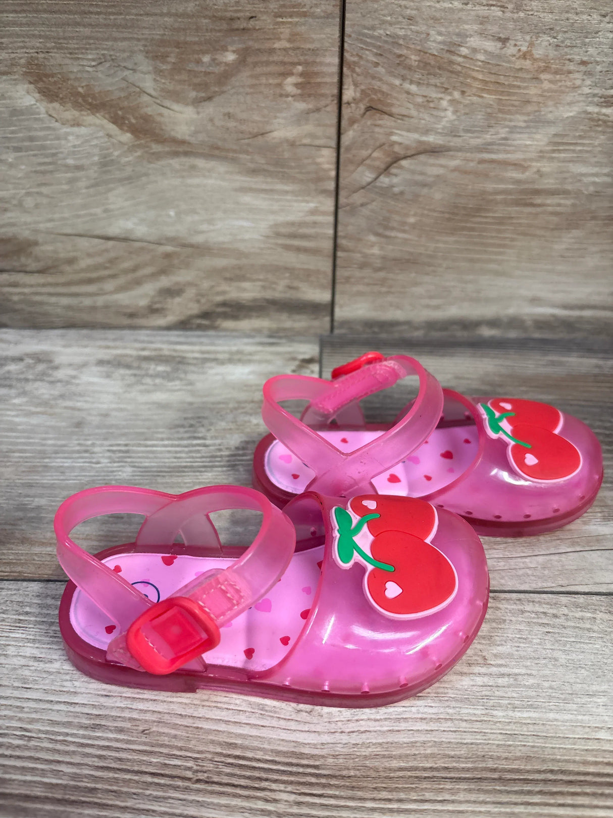 Angled view of Cat & Jack toddler pink cherry jelly sandals showing cherry appliques, heart-print insole and buckle strap on wood background