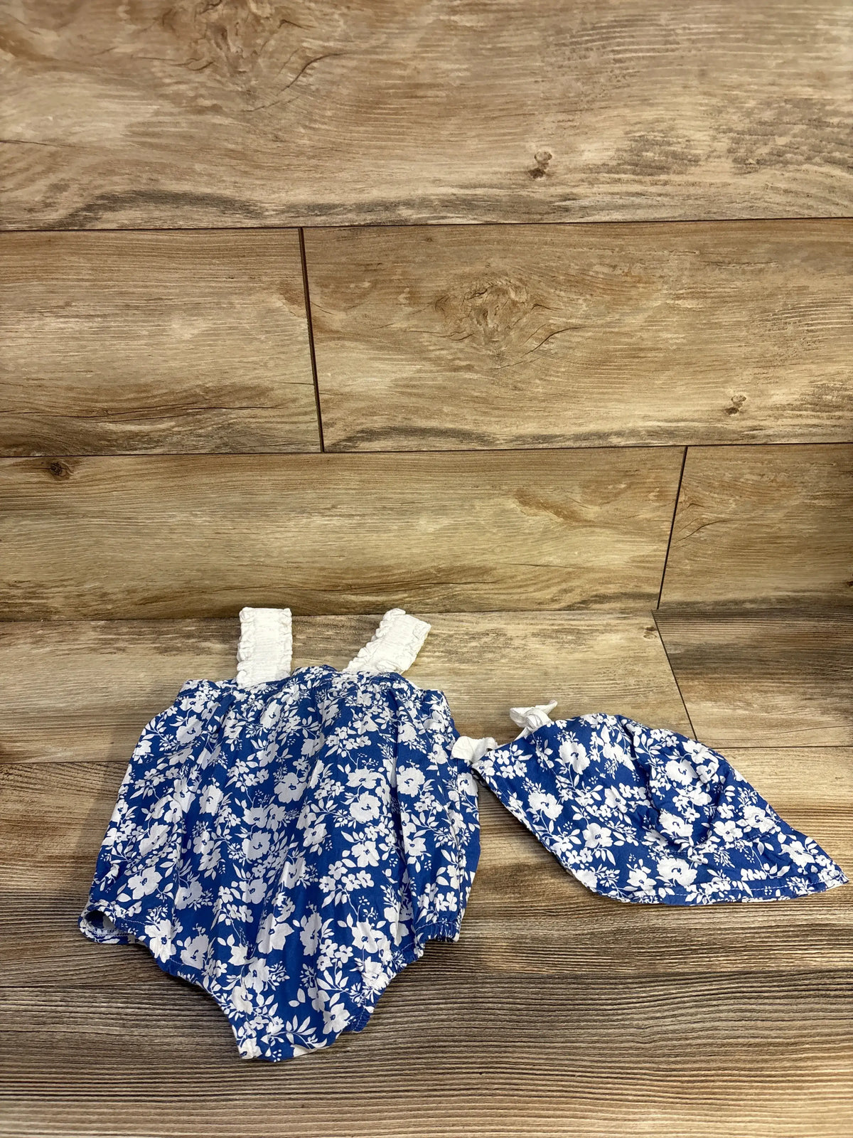 Top-down view of two-piece blue floral bubble romper and matching hat set showcasing white floral print on a wood plank background