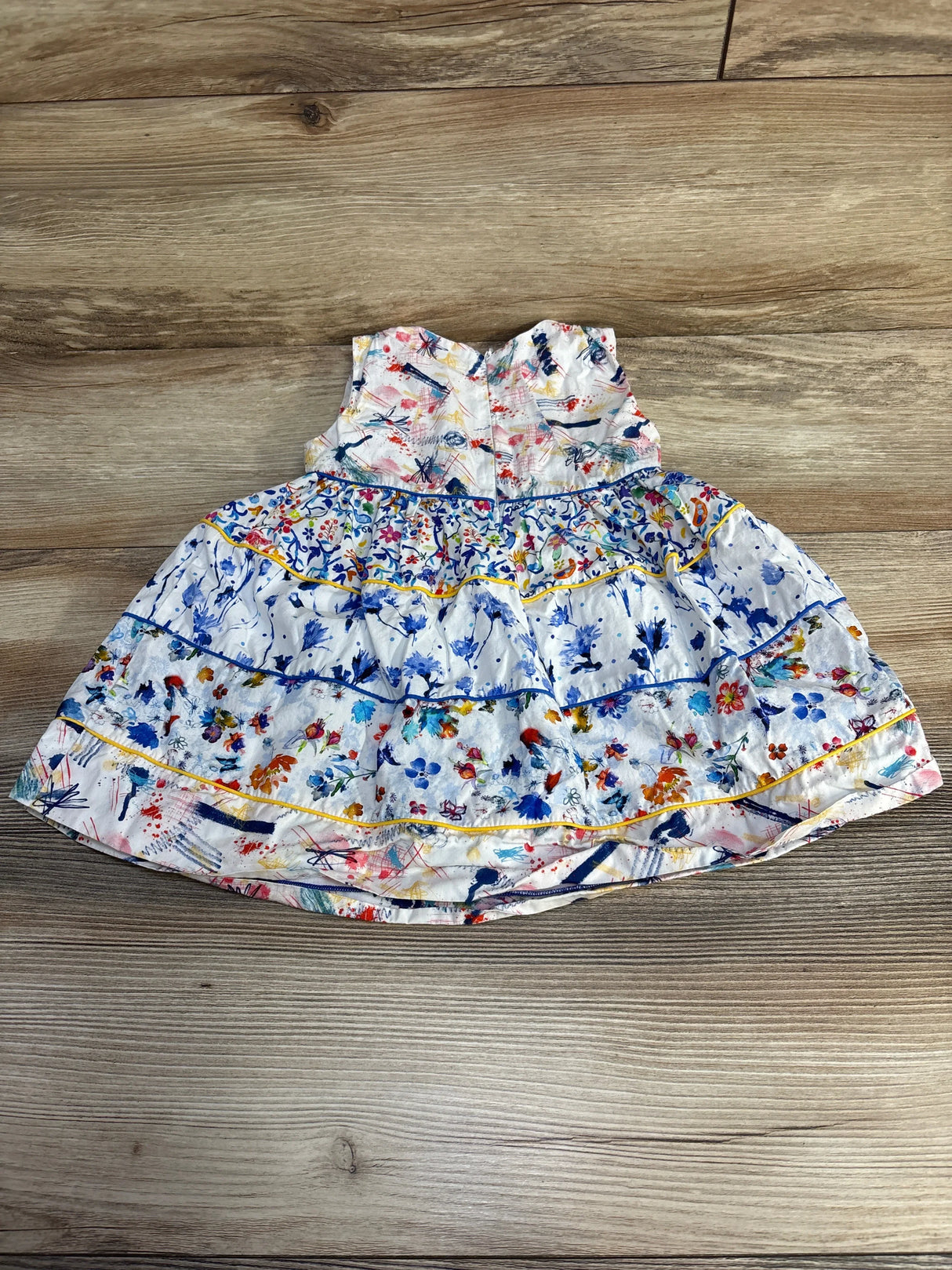 Flat lay of Fleurisse tiered toddler dress, white with multicolor paint-splatter and floral print, ruffled tiers and yellow piping on wood background