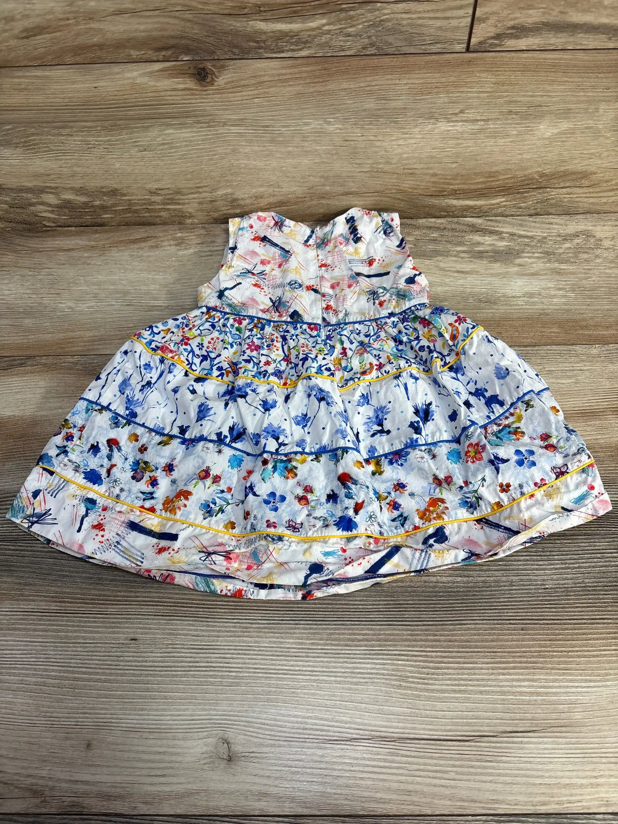 Flat lay of Fleurisse tiered toddler dress, white with multicolor paint-splatter and floral print, ruffled tiers and yellow piping on wood background