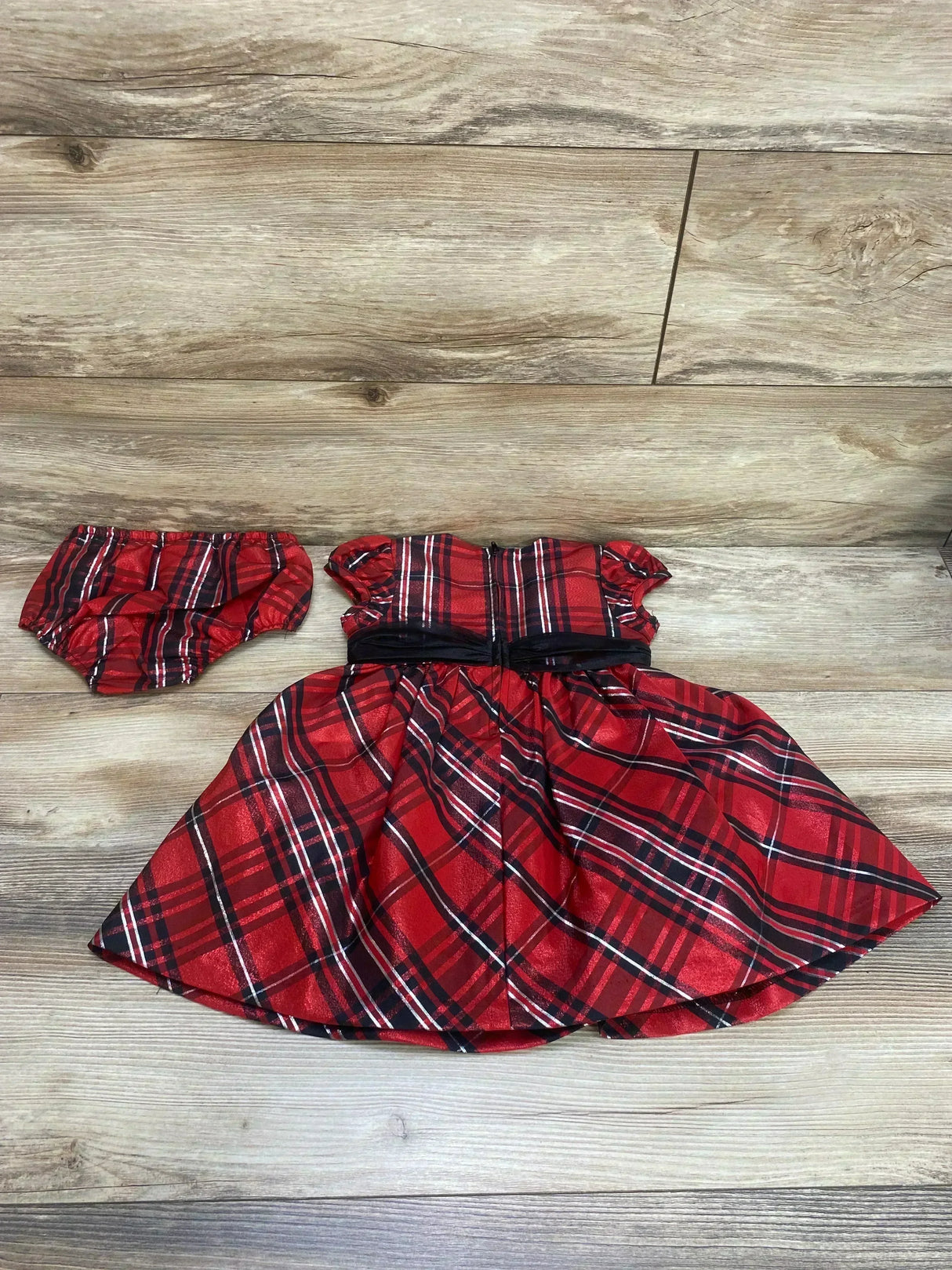 Flat lay of Bonnie Jean red plaid toddler dress and matching bloomers showing puff sleeves, black waist ribbon, and full skirt on light wood floor