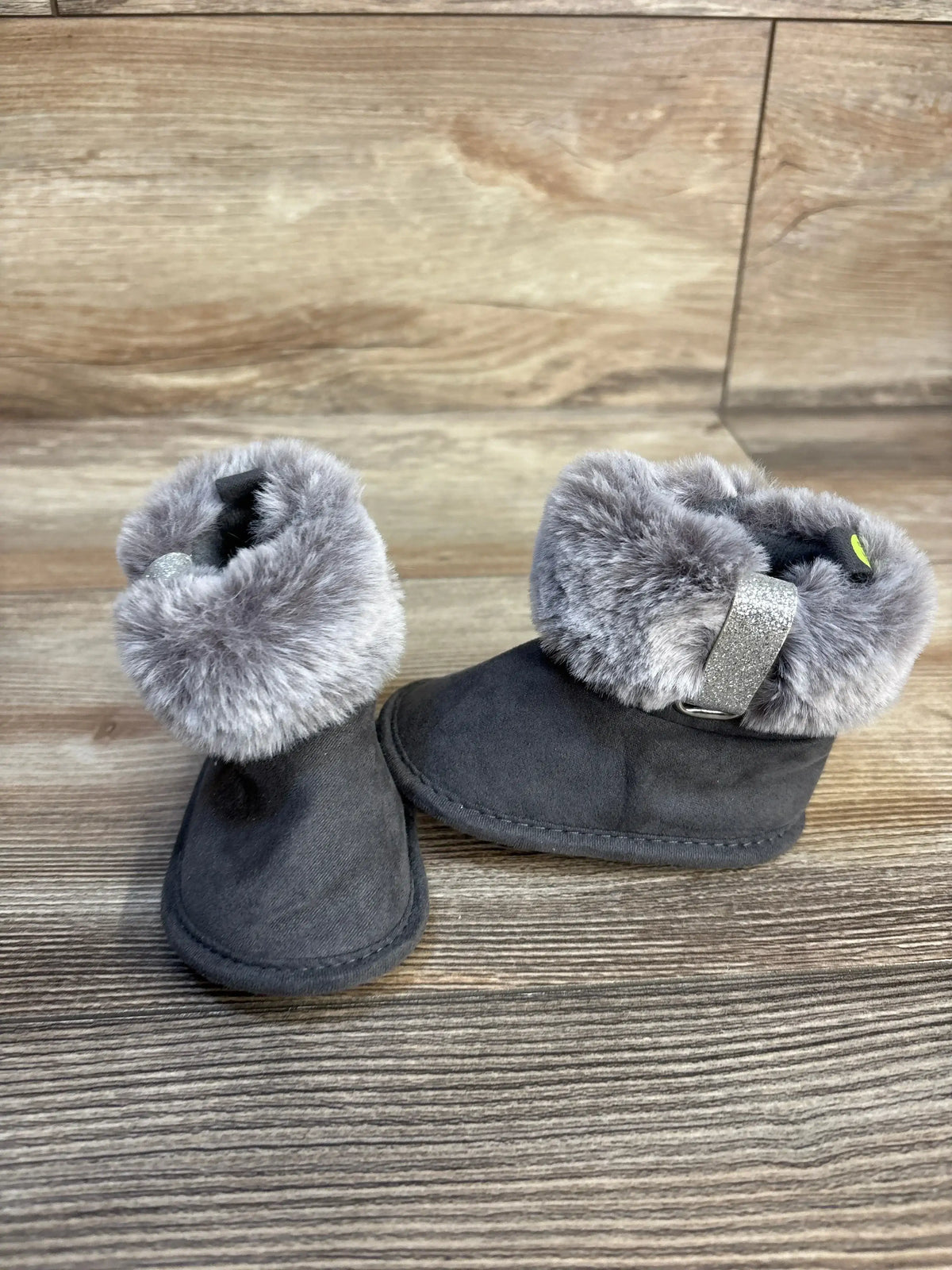 Three-quarter view of First Steps soft-sole baby boot showing grey faux-fur cuff and glitter buckle strap on wood plank floor