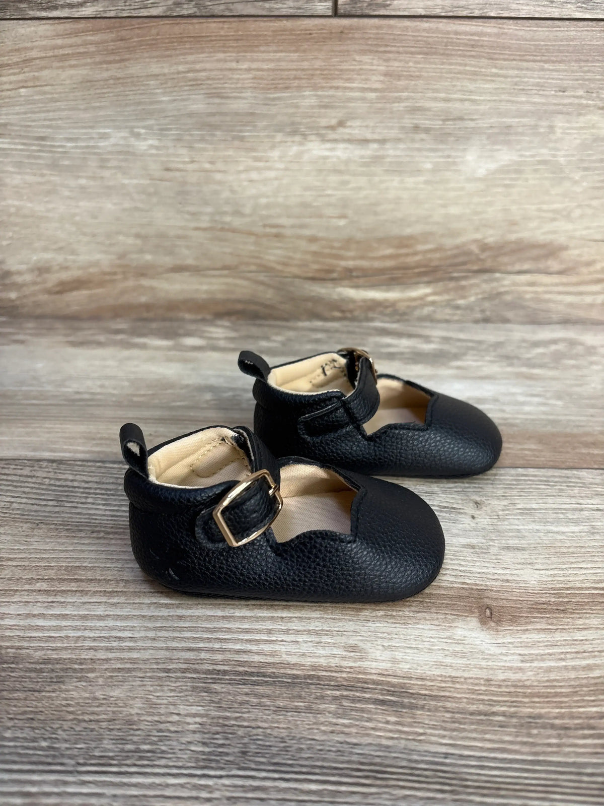Pair of black leather Me n Mommy To Be Mary Jane crib shoes with gold buckles on a wooden surface