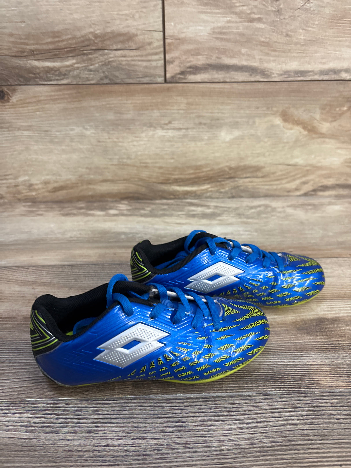 Side view of Lotto Forza II Jr soccer cleats showcasing blue upper with yellow patterned toe and white logo on wooden floor