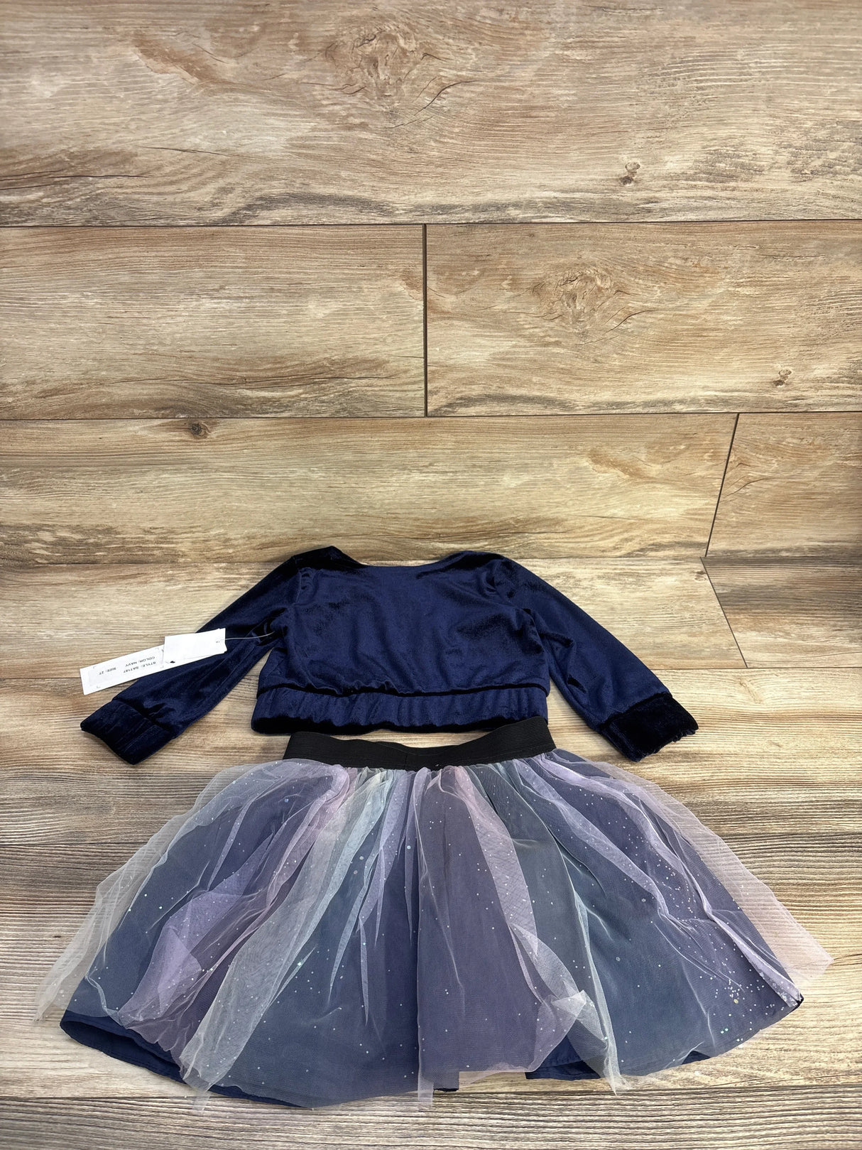 Flat lay of NWT Samara 2pc set sz 2T showcasing navy velvet bomber jacket and sparkly ombre tulle skirt on wood plank floor