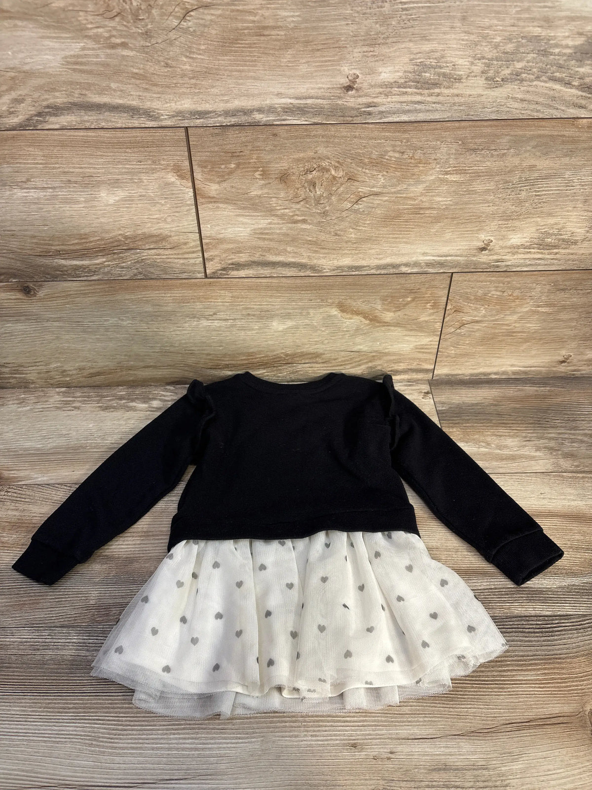 Front view of Children’s Place Paris sweatshirt dress showcasing black long sleeves and a white heart-patterned mesh skirt on a wood floor