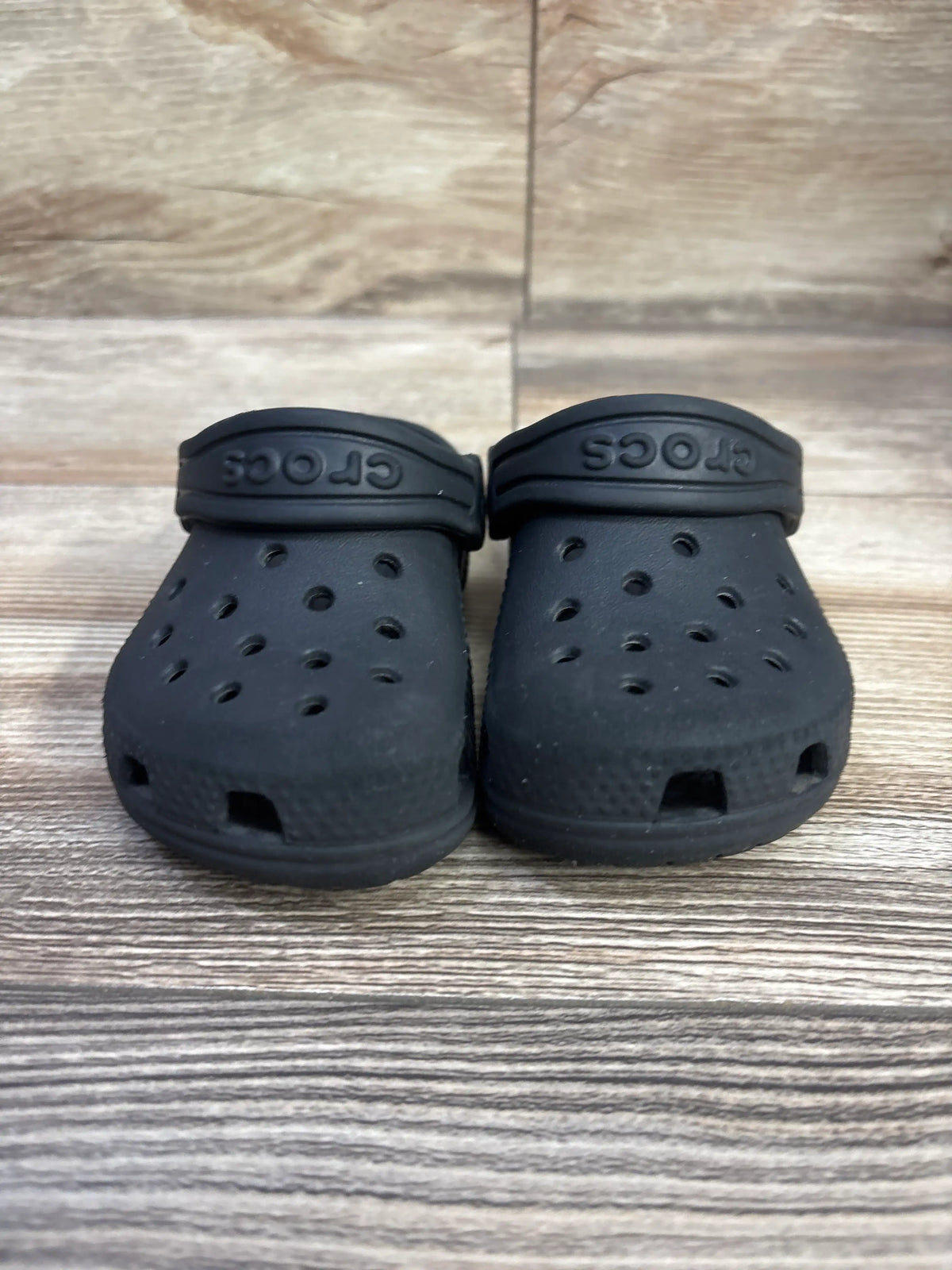 Front view of black Crocs Classic Clogs in size 6C showcasing ventilation holes and heel straps with a wood-patterned background