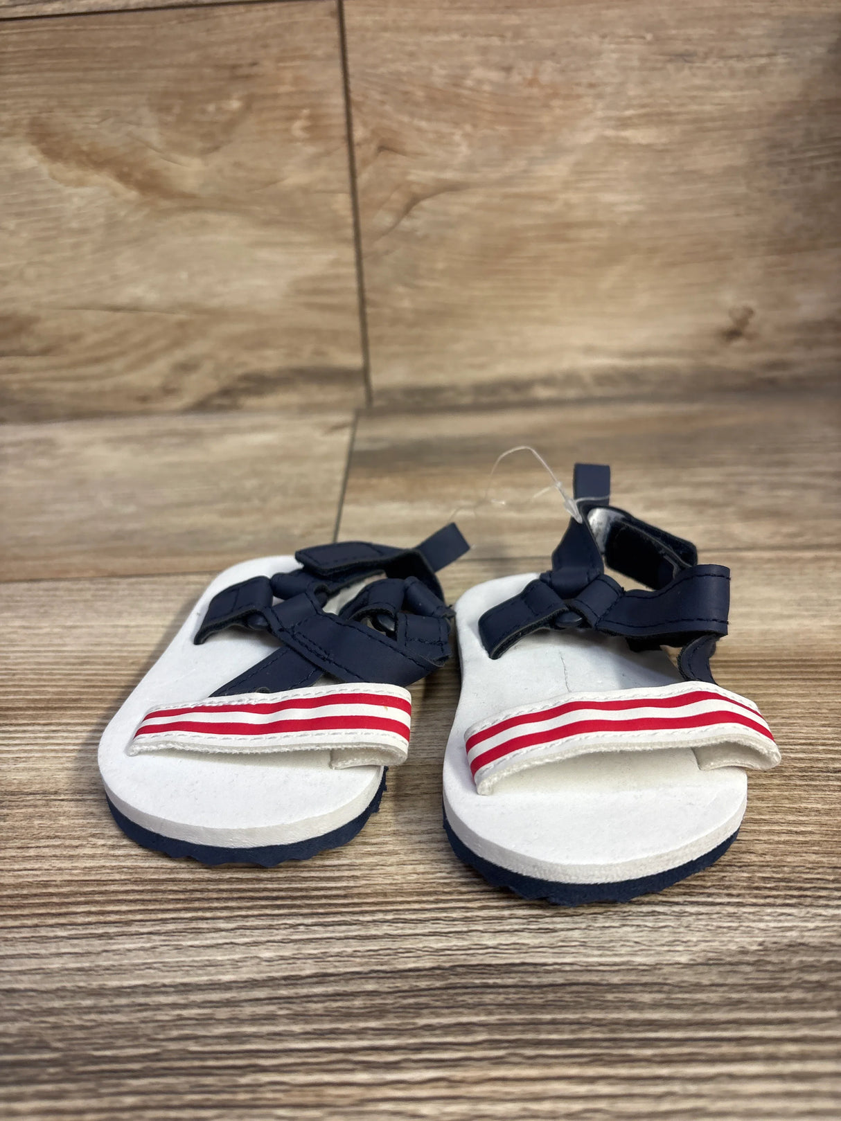 Front view of Child Of Mine baby sandals, white footbed with navy straps and red-and-white striped front strap, size 3-6 months on wood background