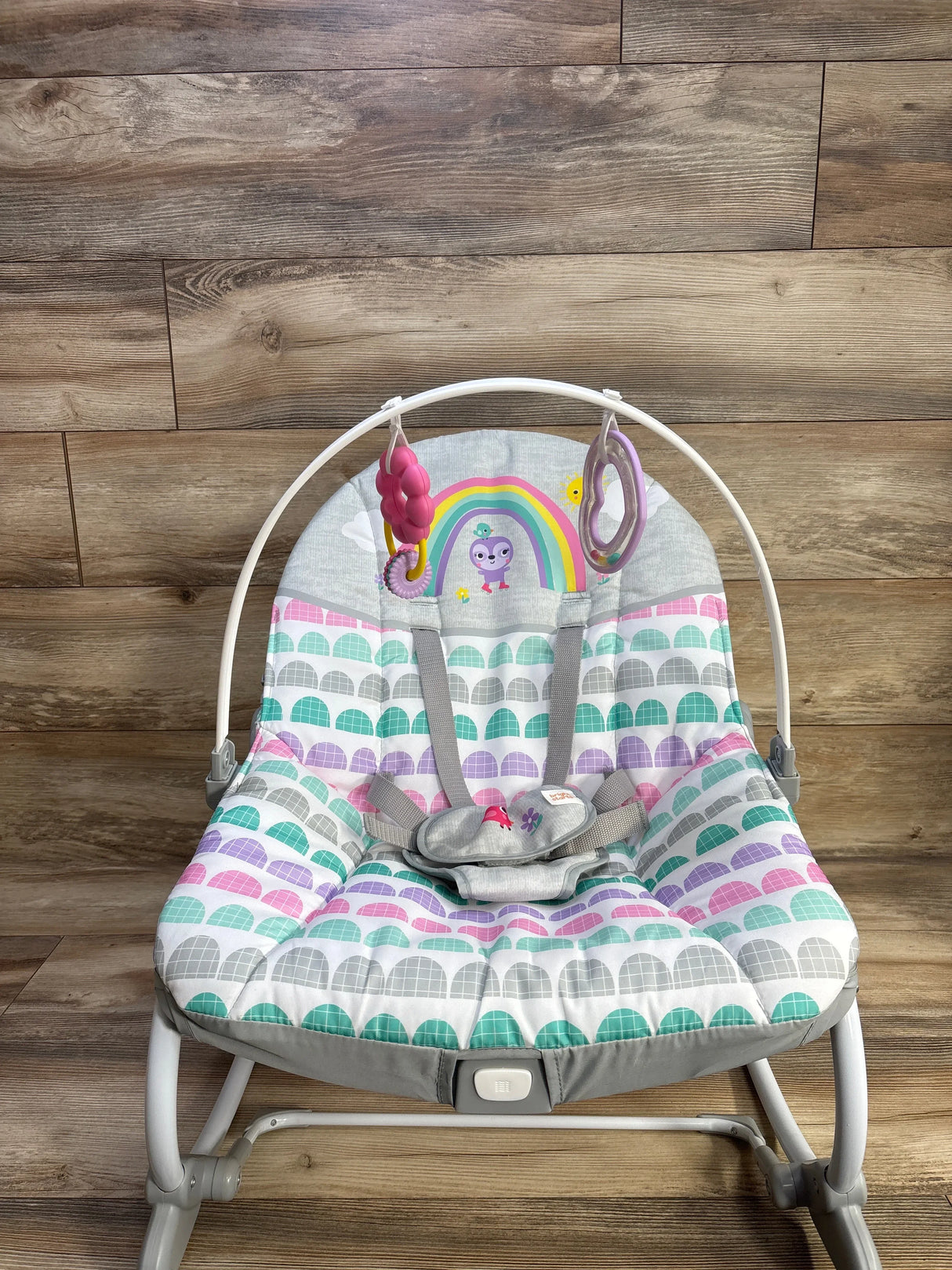 Front view of Bright Starts Rosy Rainbow rocker with pastel scallop seat, toy bar with two hanging toys and gray harness