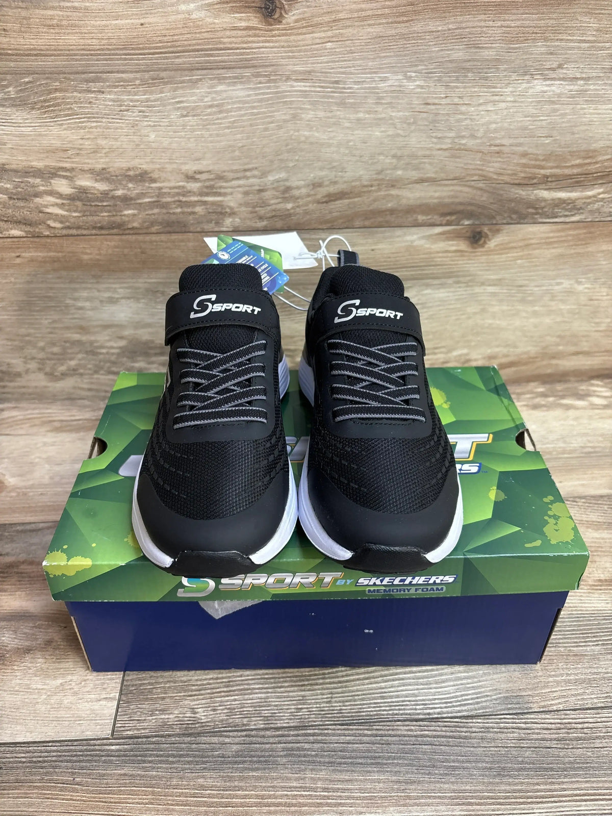 Black S Sport by Skechers sneakers with white soles on a branded shoebox, wood floor background