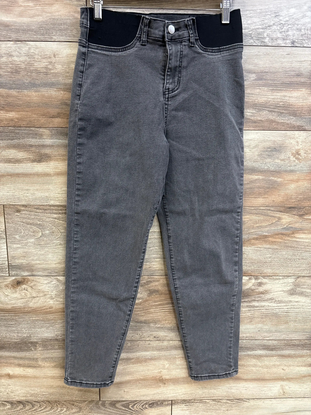 Front view of grey maternity jeans with black elastic belly panel, button closure and tapered legs on wood background