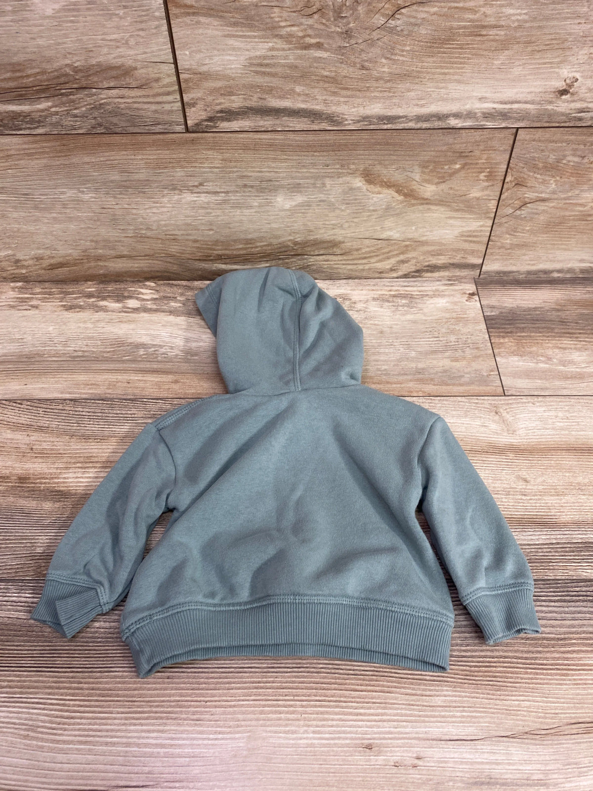 Back view of Little Co. Be The Good green pullover sweatshirt showcasing its hood and ribbed cuffs on a wood-look floor