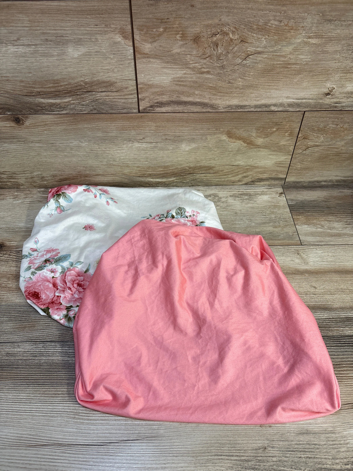 Top-down view of two fitted bassinet sheets showcasing white floral and solid pink designs and elasticized edges on a wood plank background