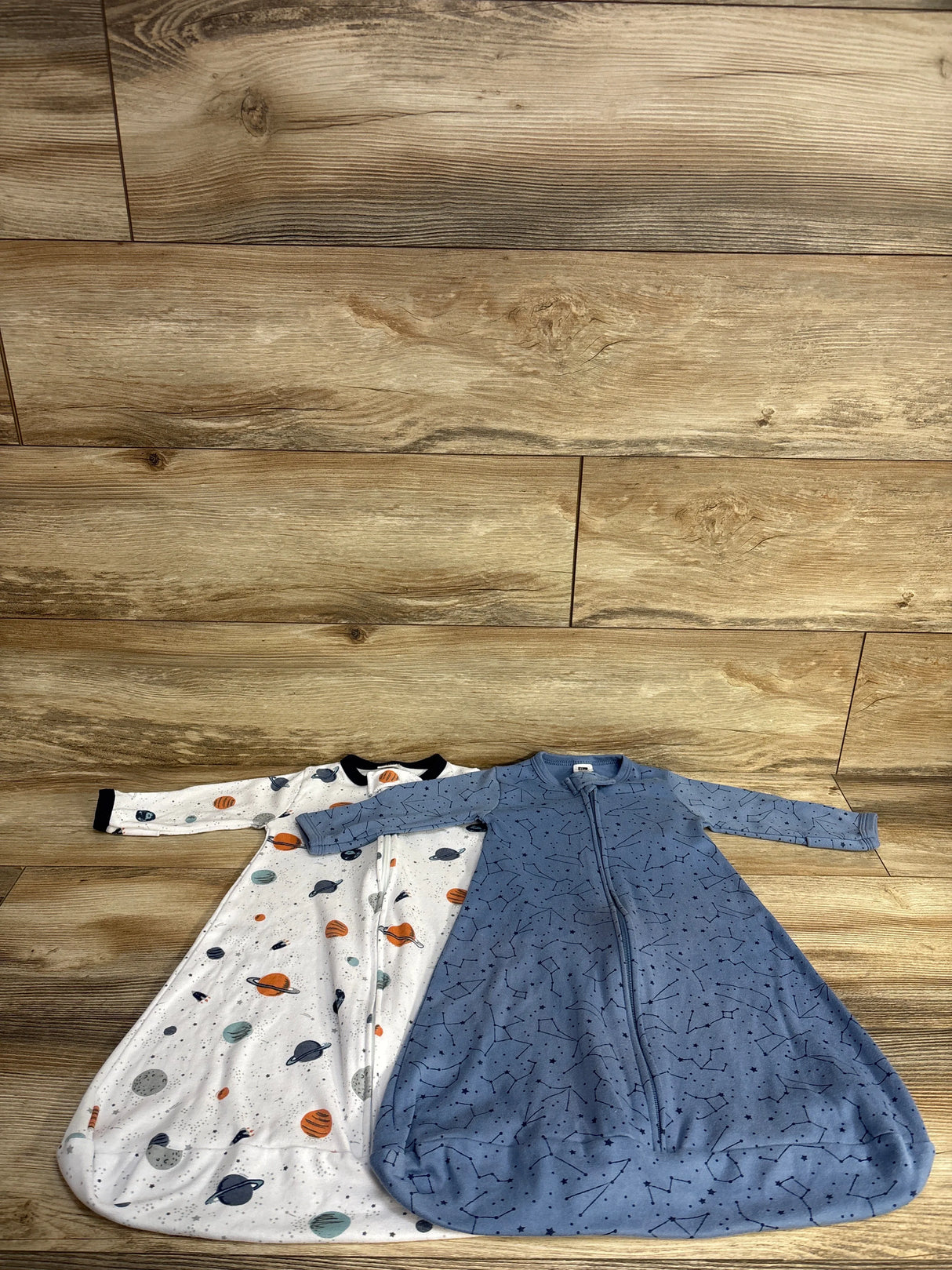 Two baby sleep sacks on wooden floor, one white with planets and one blue with constellations pattern