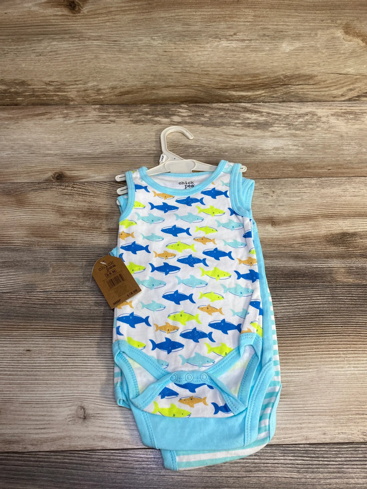 Top view of 3-pack Chick Pea tank bodysuits showing blue-trimmed shark-print front bodysuit on hanger on wood background