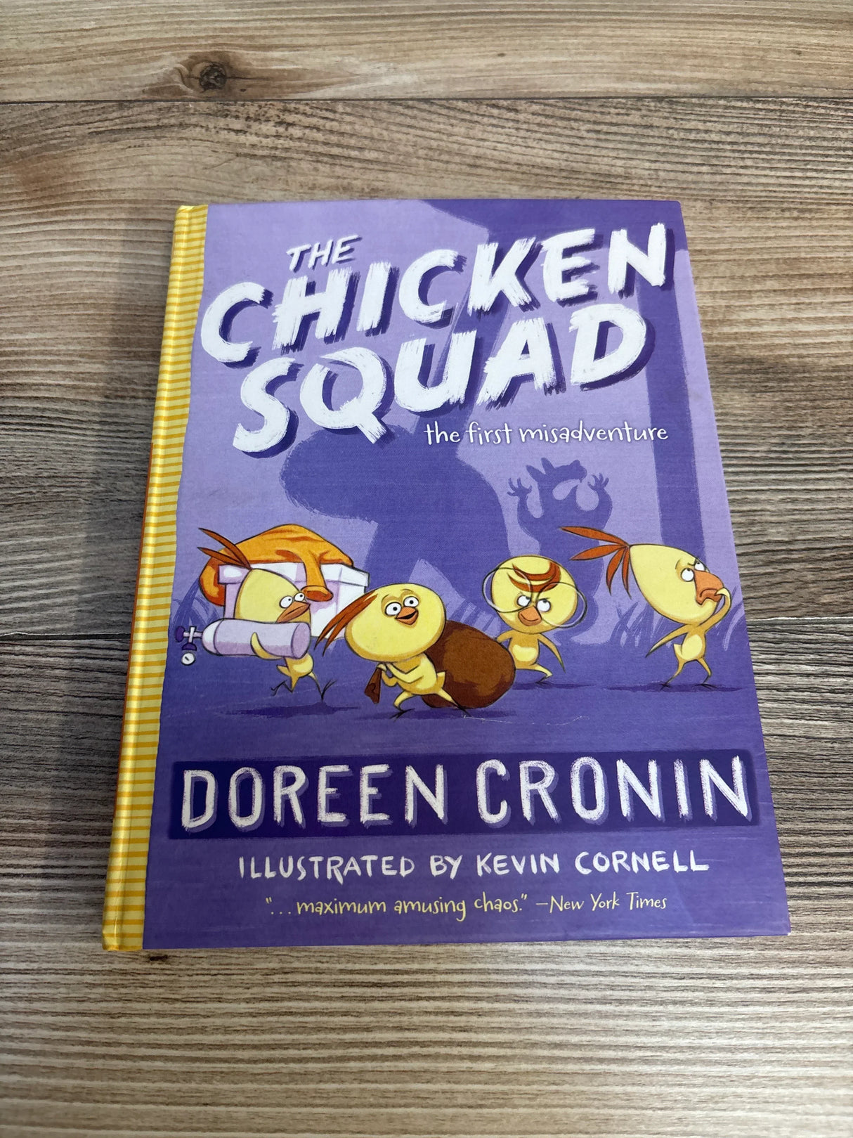 Top-down view of hardcover The Chicken Squad: The First Misadventure showing purple illustrated cover with cartoon chicks and yellow-striped spine on wood table