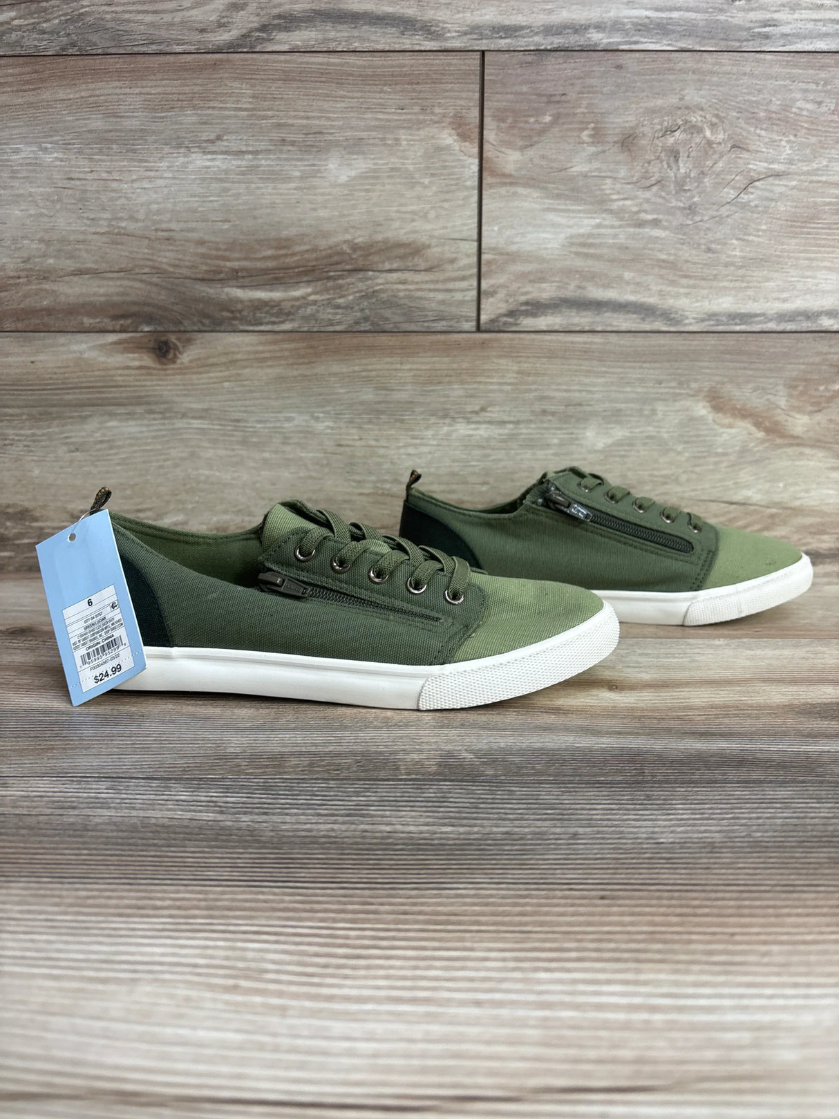 Pair of green casual kids canvas sneakers with white soles and side zippers on wood floor background, size tag attached