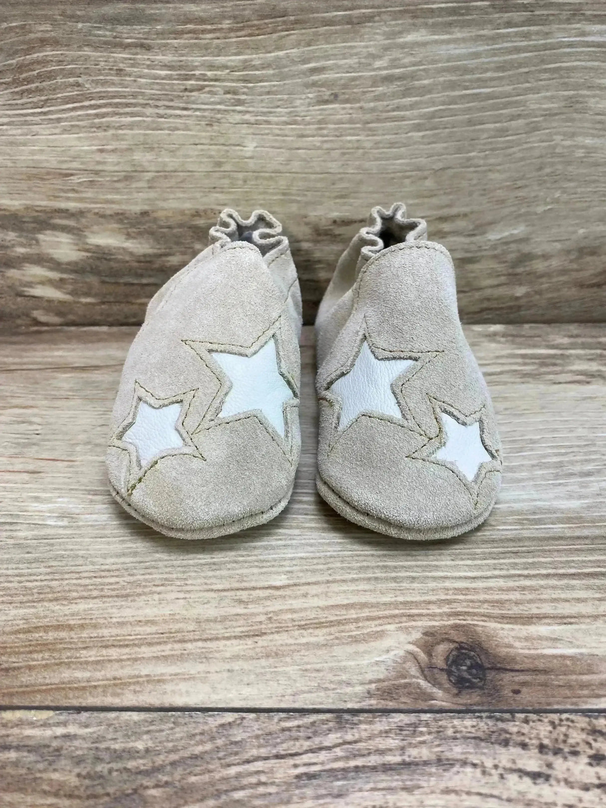 Front view of Minnetonka tan suede infant booties showing white star appliques and ruffled elastic ankles on wood background