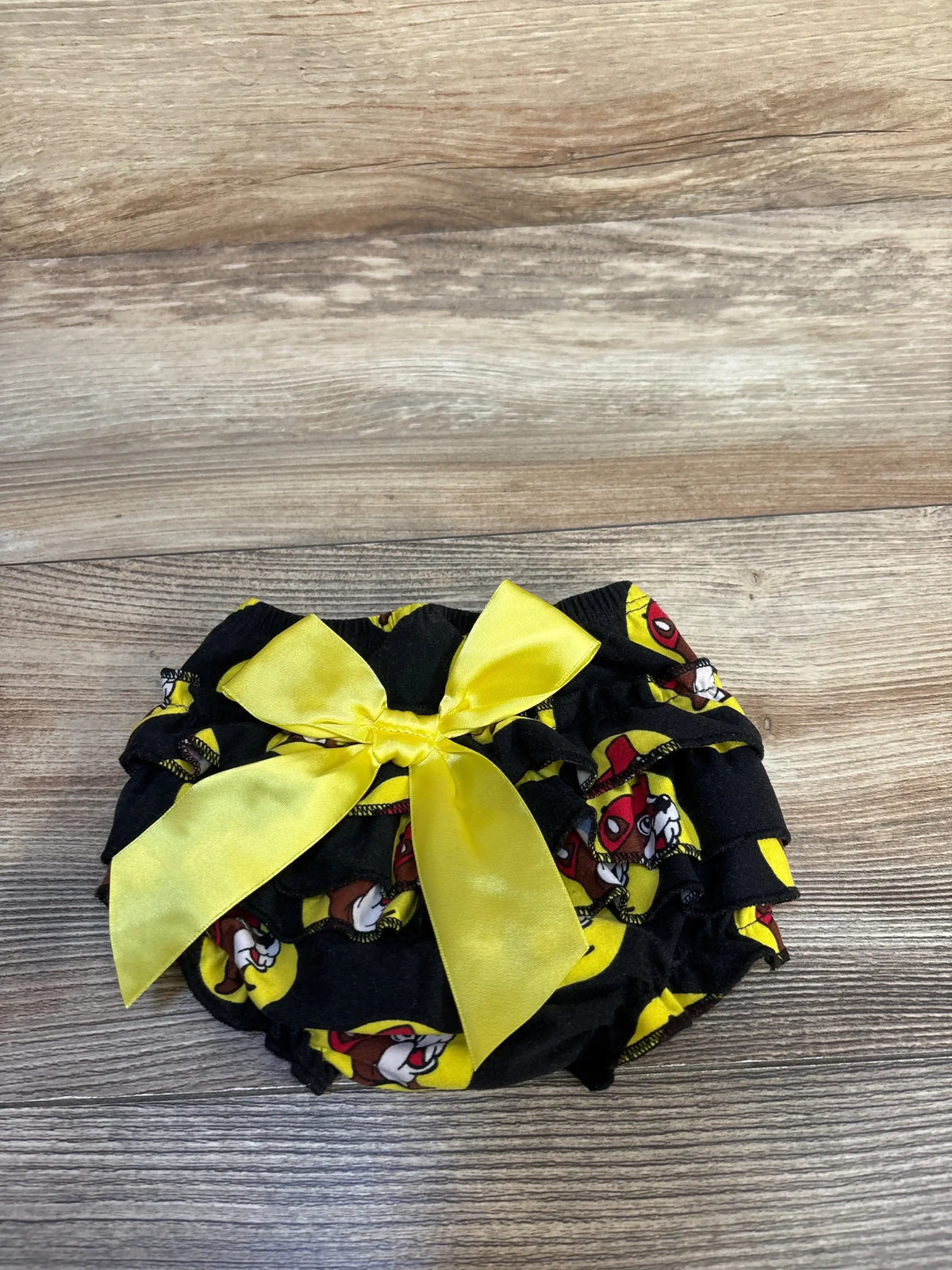 Black baby ruffle diaper cover with cartoon print and large yellow satin bow, on wood surface.