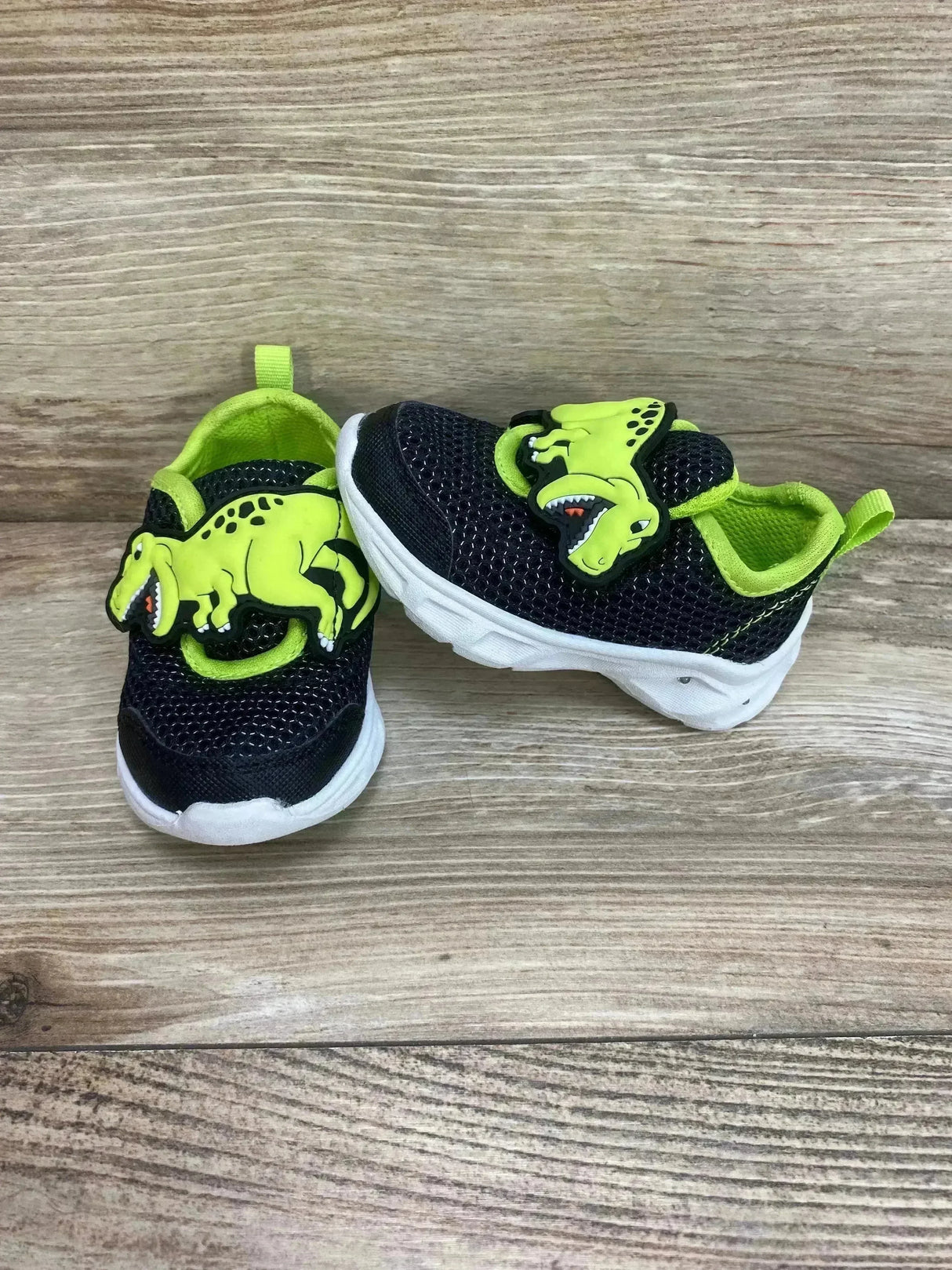 Angled view of Carter's toddler dinosaur light-up shoe, black mesh with neon green dinosaur applique and white sole on wood background
