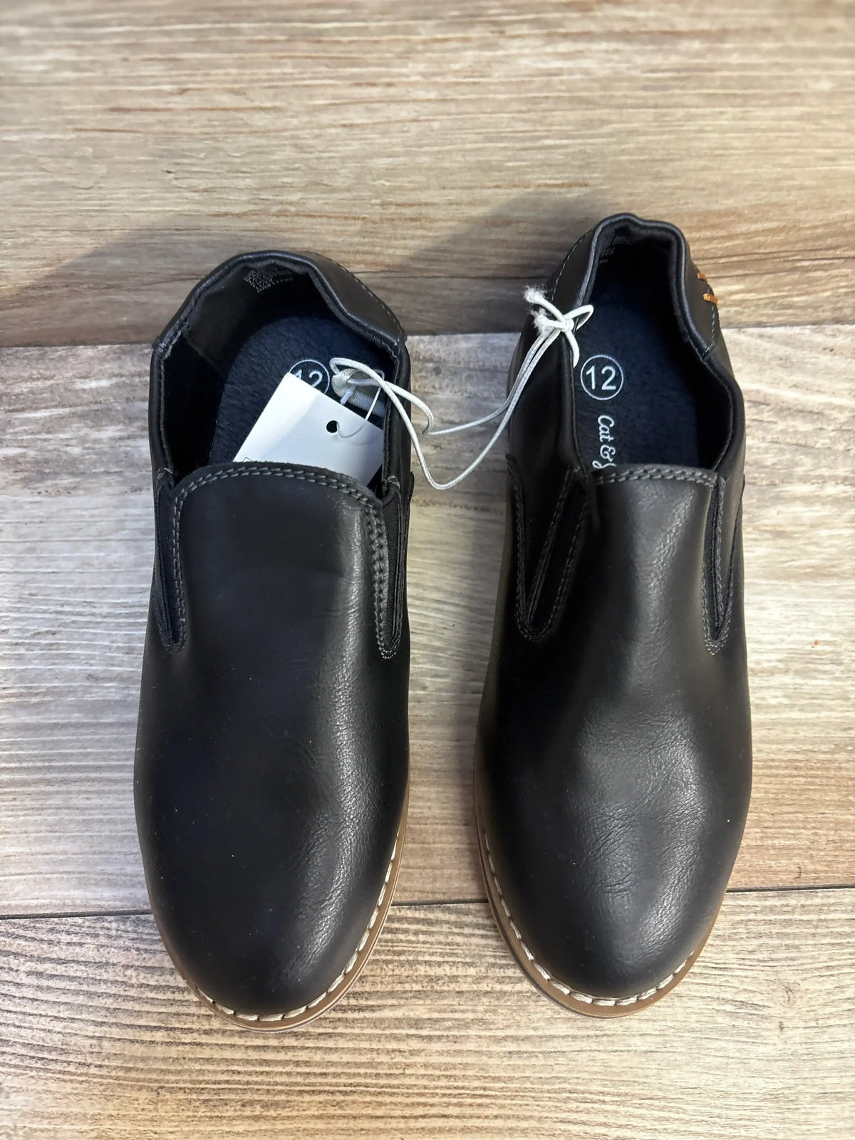 Pair of black slip-on kids shoes with tags, size 12, on wooden floor