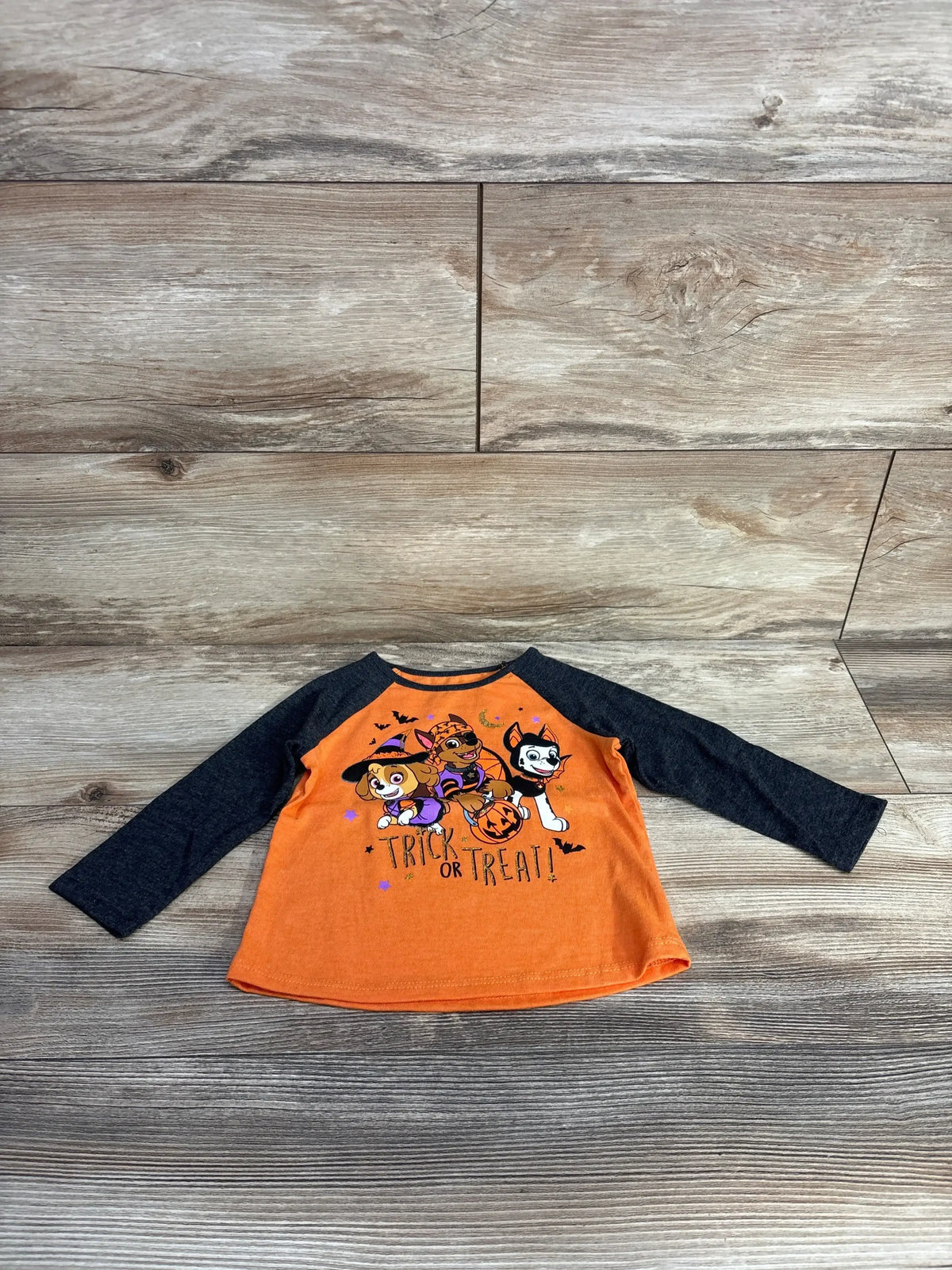 Front view of Jumping Beans Paw Patrol Trick or Treat shirt with orange body, black sleeves, and graphic on wood floor