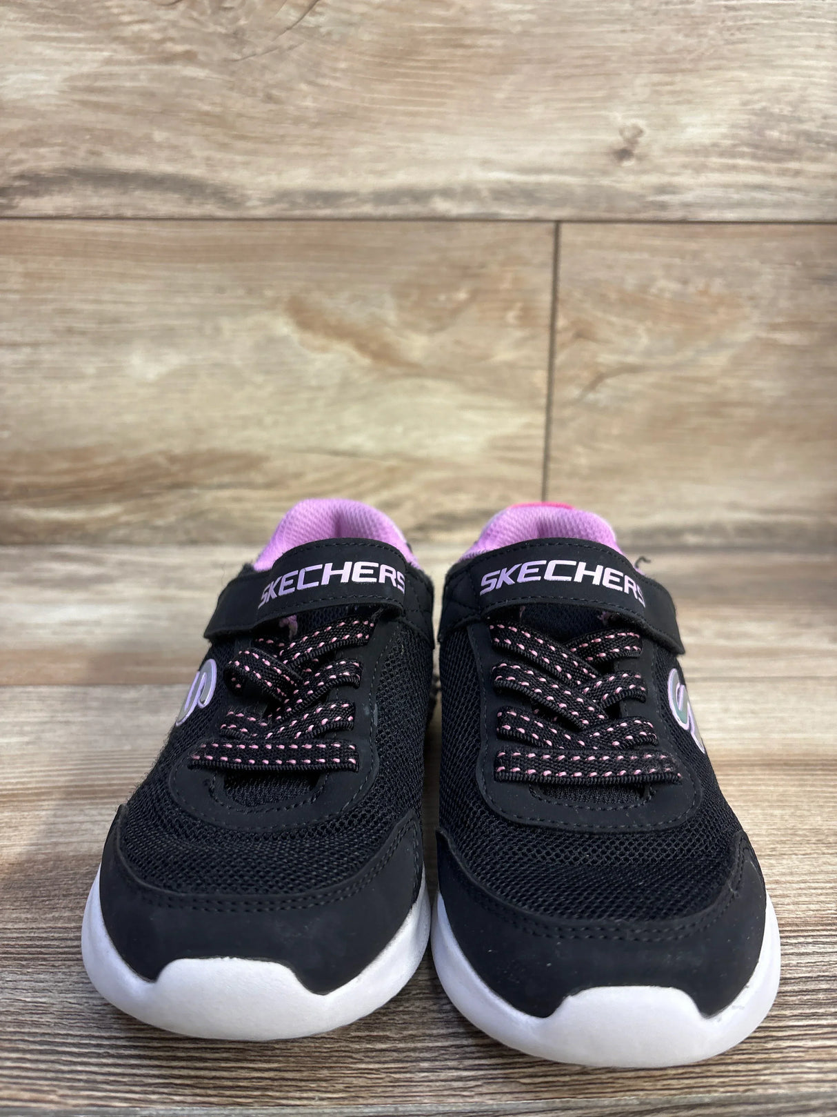 Front view of black Skechers Ultra Flex Floral toddler sneakers with pink lining, white soles, and Velcro strap on wood floor