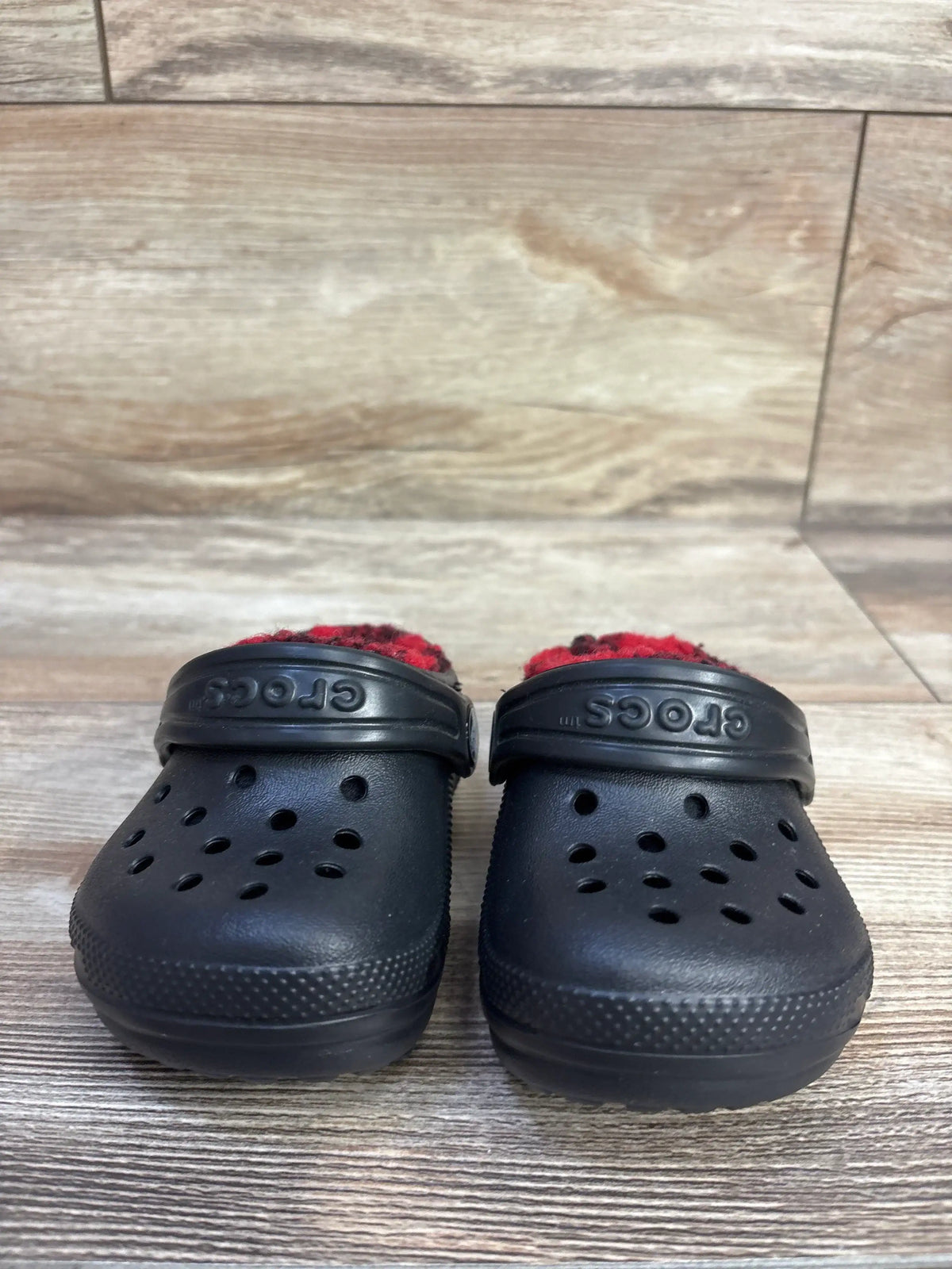 Front view of black Crocs Classic Lined toddler clogs showing red buffalo-check fleece lining on wood floor