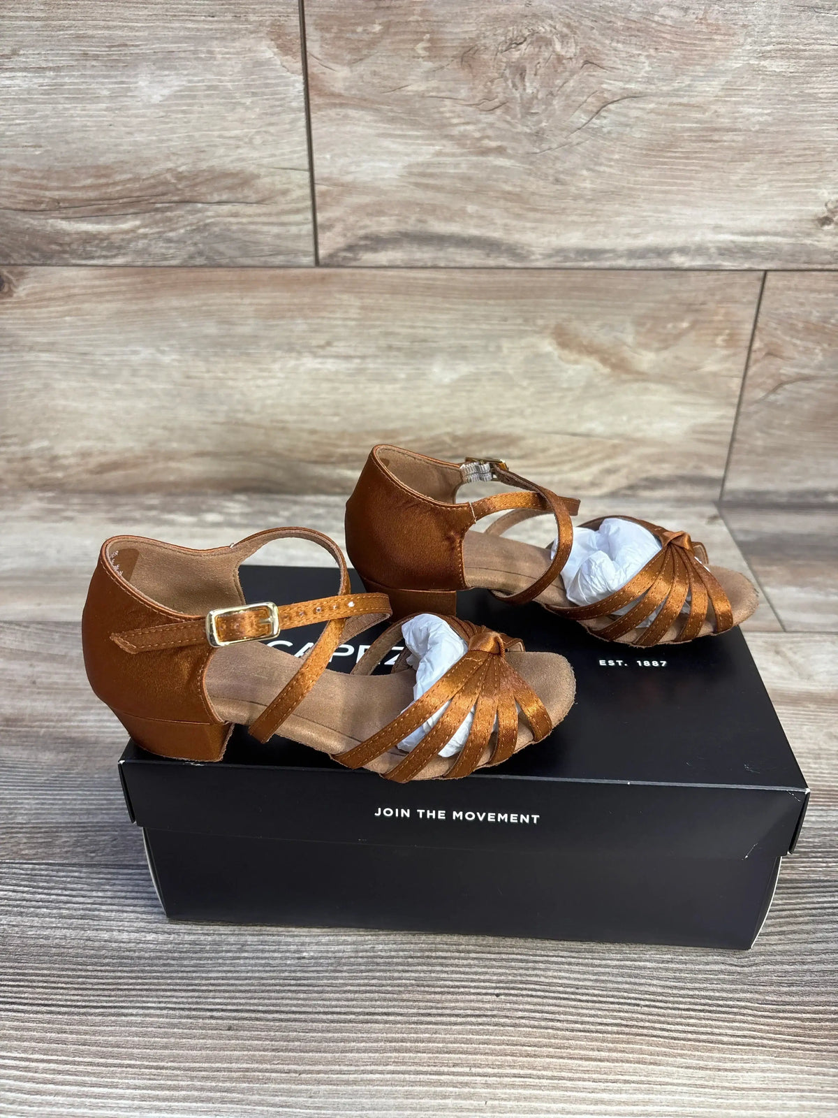 Three-quarter view of Capezio Valentina ballroom flats in cinnamon showing crisscross straps, ankle buckle and low heel on a black shoebox