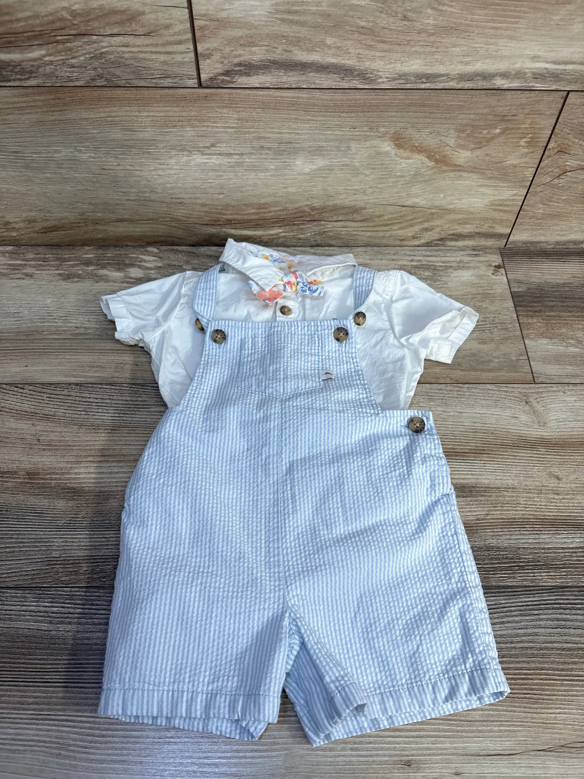 Flat lay of Tommy Bahama baby outfit showing white polo bodysuit, blue striped shortalls, and bowtie on wood flooring