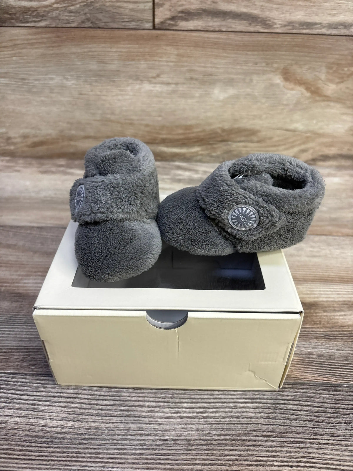 Three-quarter view of Ugg Bixbee baby booties showcasing grey plush fleece and strap with UGG button, on cream box and wood background